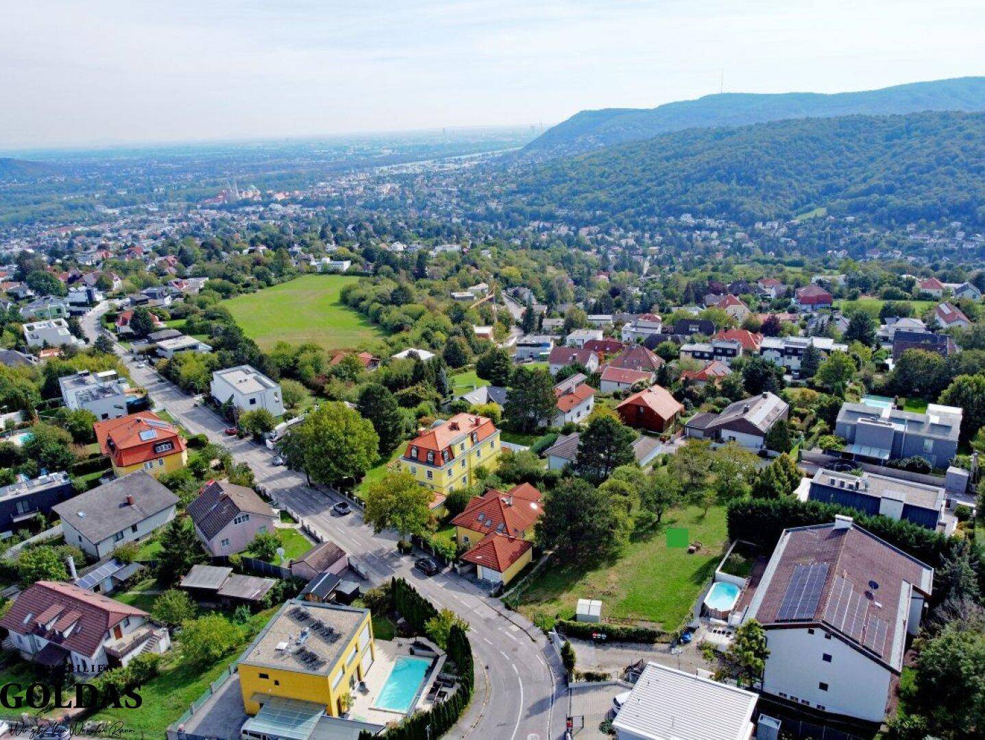 Panoramablick aus der Luft auf ein Wohngebiet mit grünen Hügeln und einem freien Baugrundstück.