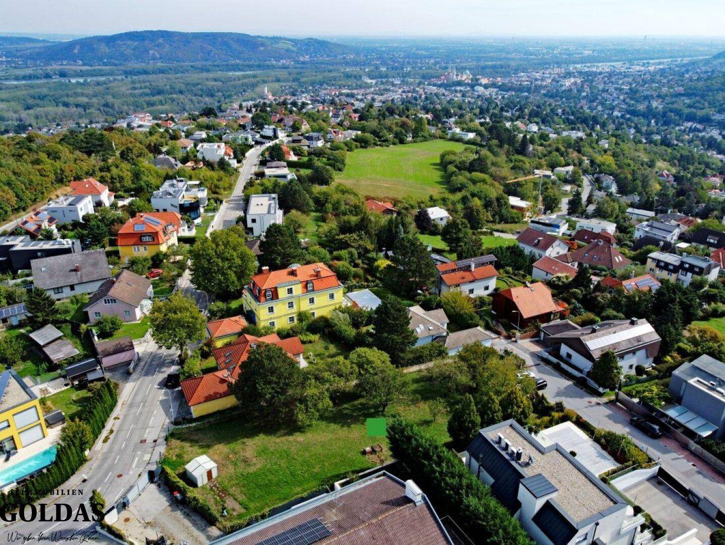 Weitläufige Luftaufnahme eines Wohngebiets mit viel Grün und einem freien Grundstück.