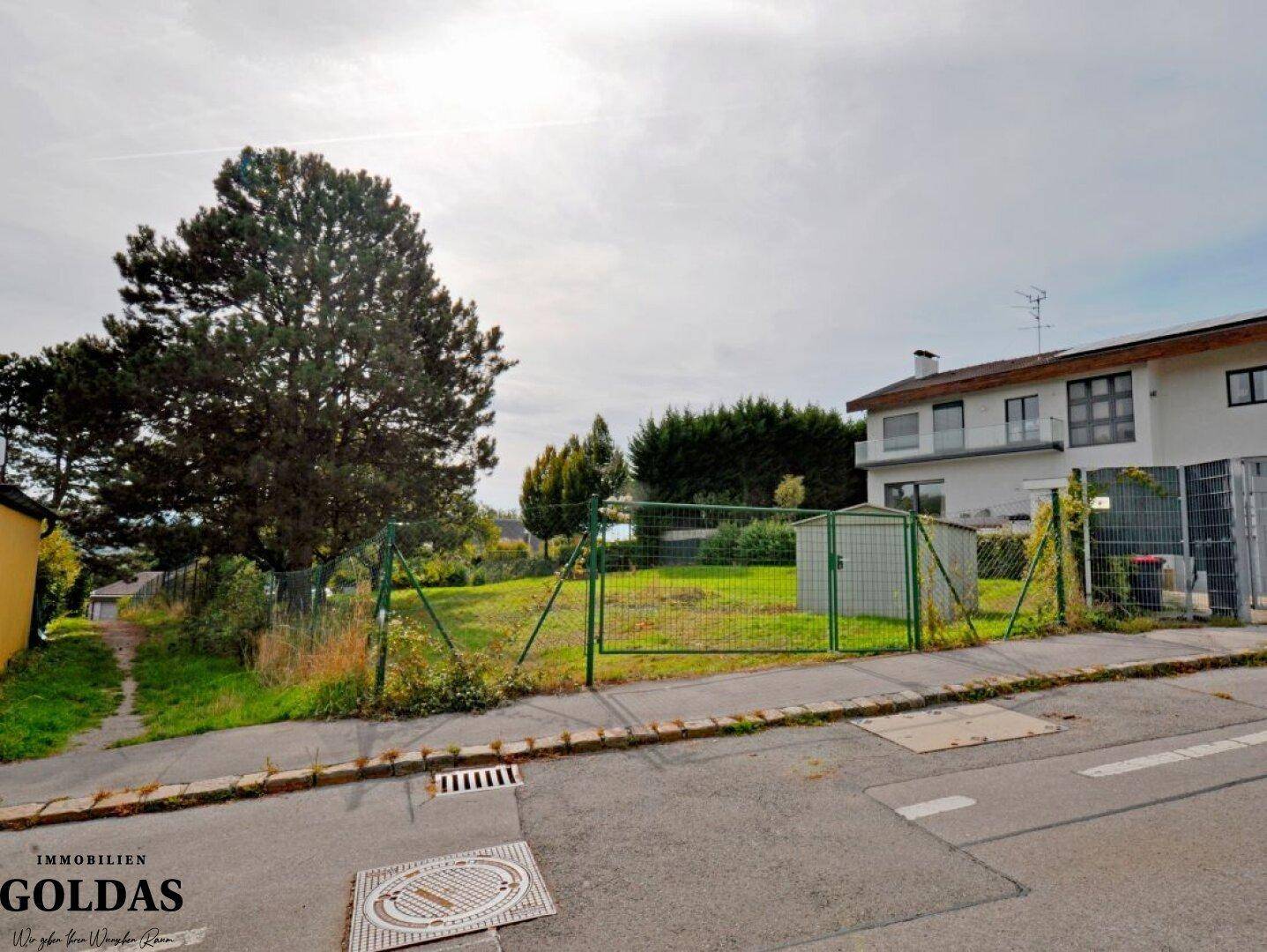Blick von der Straße auf ein eingezäuntes Baugrundstück mit einem Gartenschuppen.