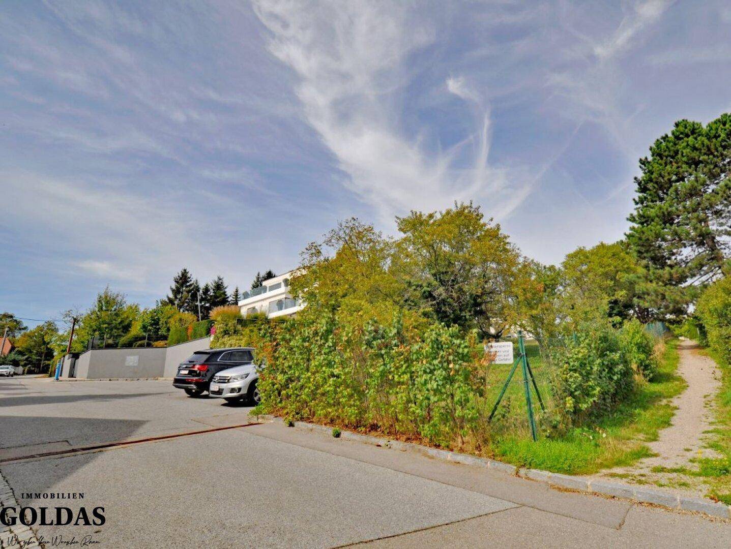 Straßenansicht mit Parkplätzen und einer grünen Hecke, die das Grundstück begrenzt.