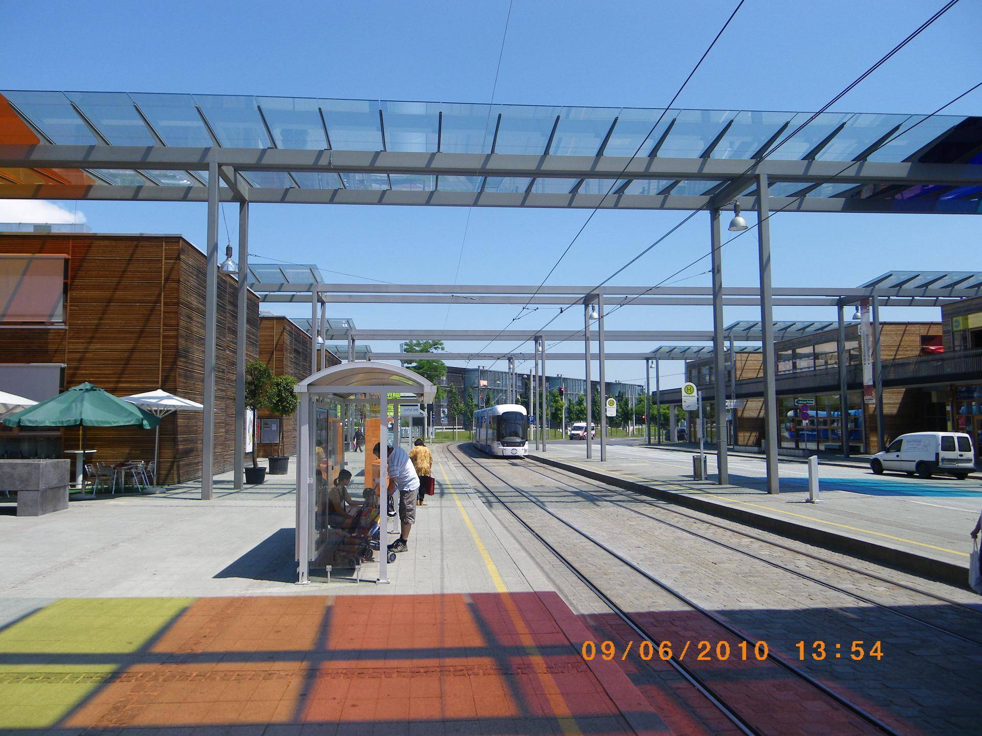 Eine Straßenbahnhaltestelle an einem sonnigen Tag mit modernen Gebäuden und Gleisen in einer städtischen Umgebung.