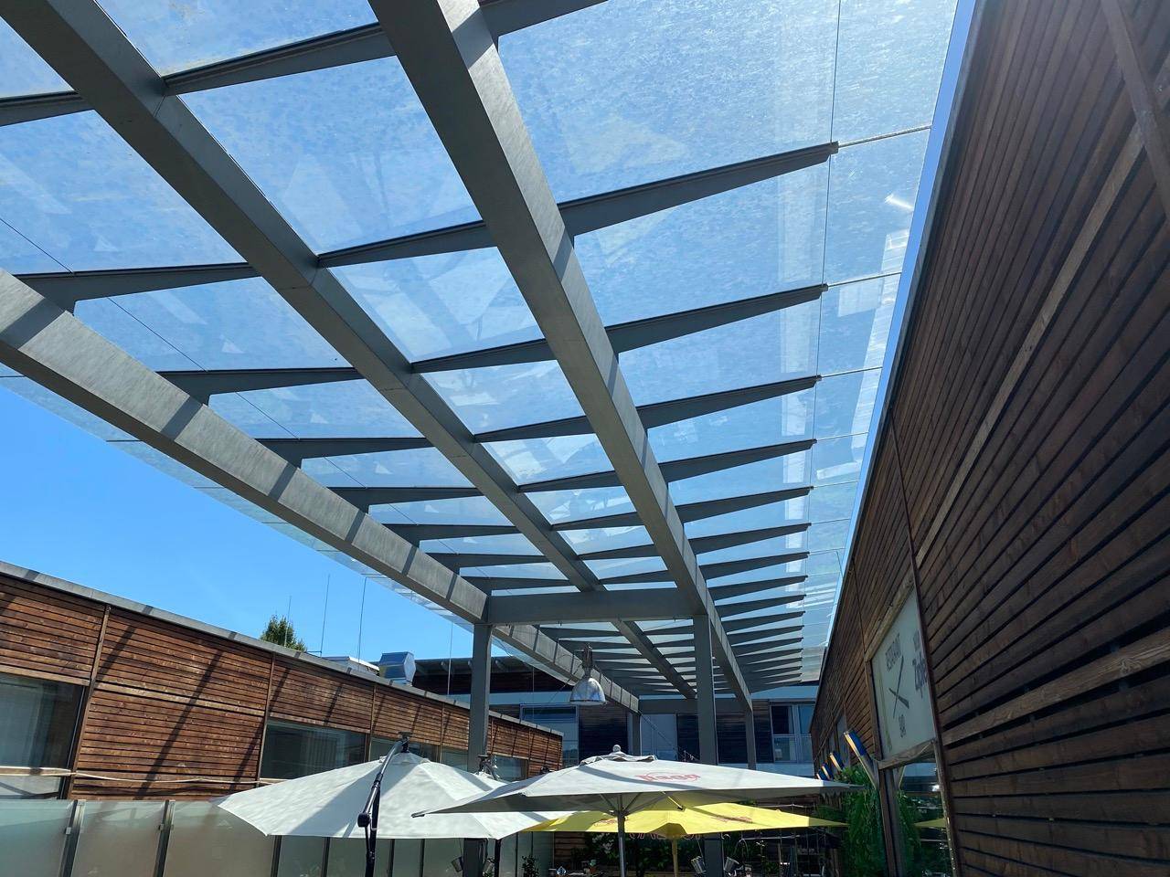 Moderne Glasdachkonstruktion über einem Außenbereich mit Sonnenschirmen und Holzfassaden unter blauem Himmel.