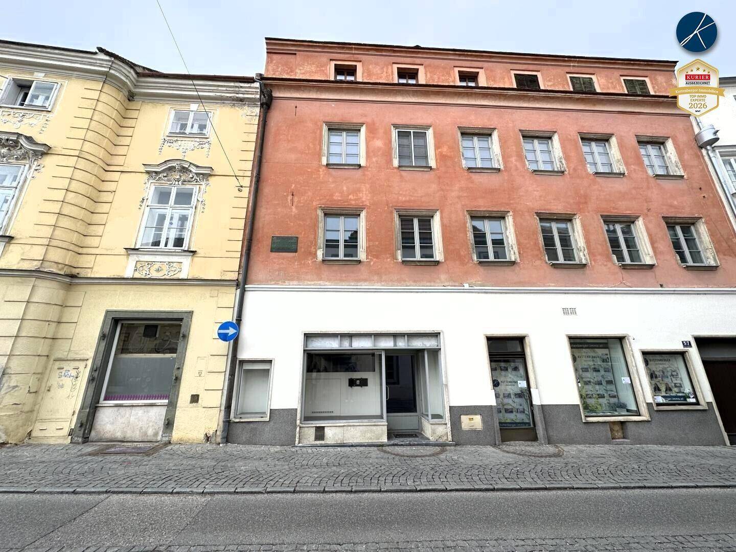 Straßenansicht zweier historischer Gebäude mit Ladenfronten im Erdgeschoss und Wohnungen darüber, in städtischer Umgebung.