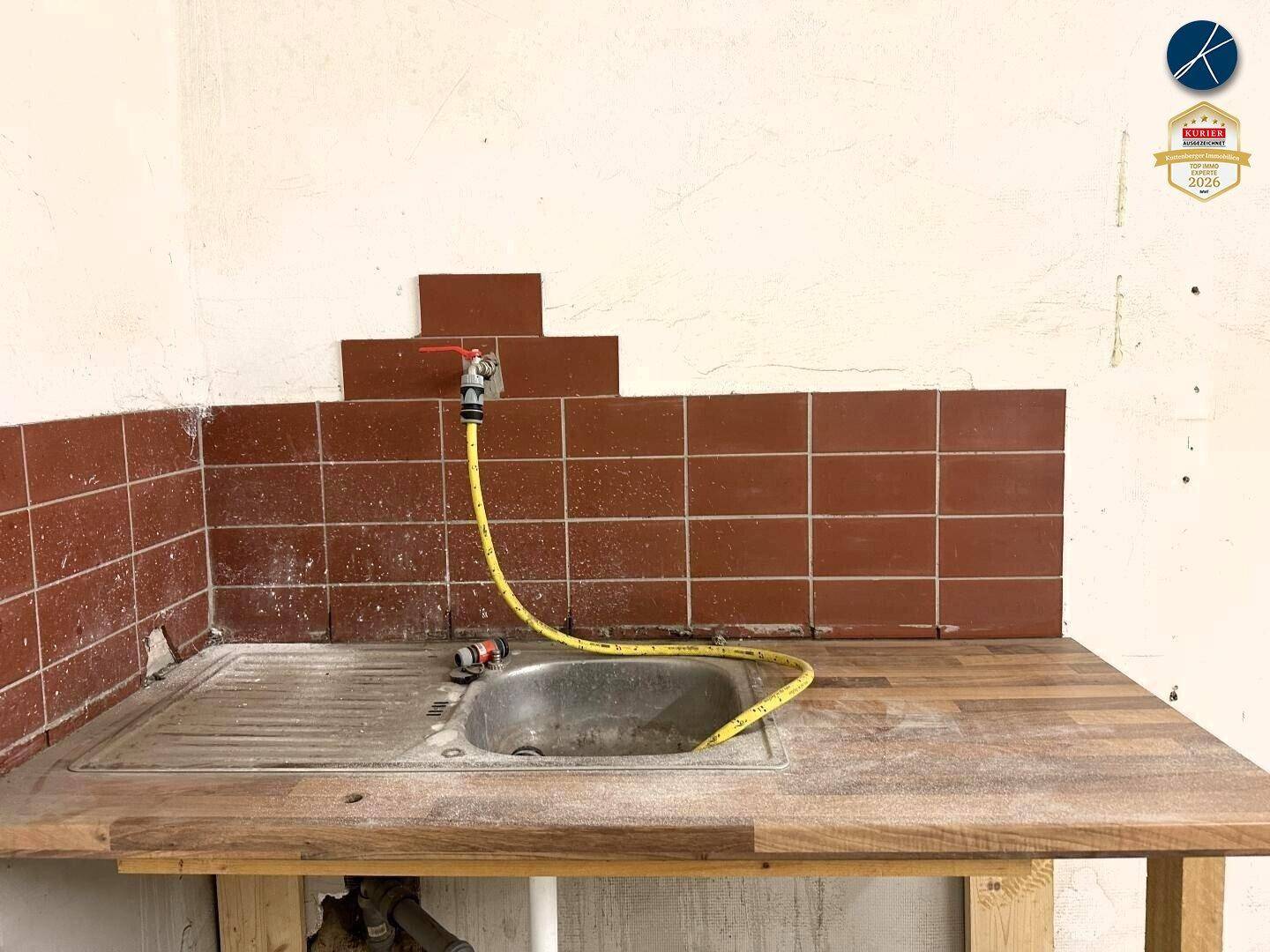 Detailansicht einer einfachen Werkbank mit Spülbecken, Wasserhahn und braunen Wandfliesen in einem Nebenraum.