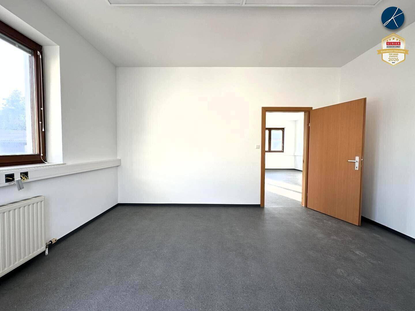 Heller Büroraum mit Fenster und offener Tür, die den Blick in einen weiteren Raum freigibt.