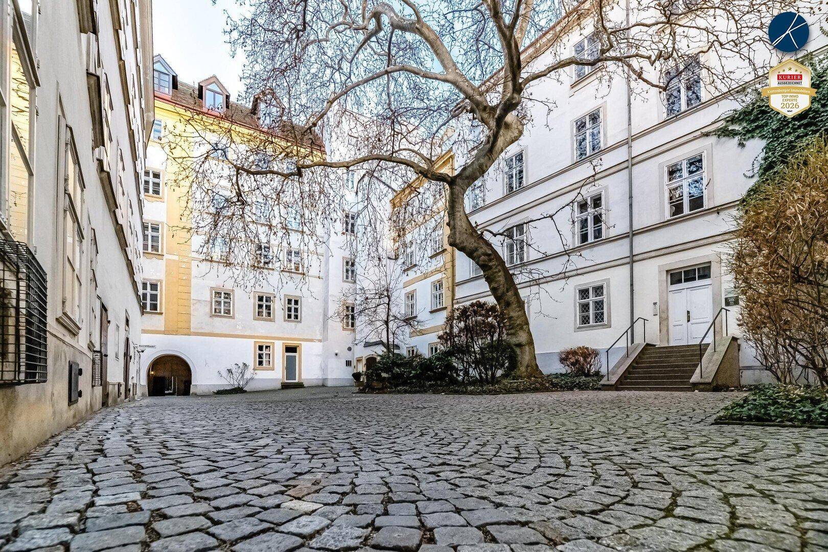 Malerischer Innenhof mit Kopfsteinpflaster und einem großen Baum, umgeben von historischen Gebäuden.