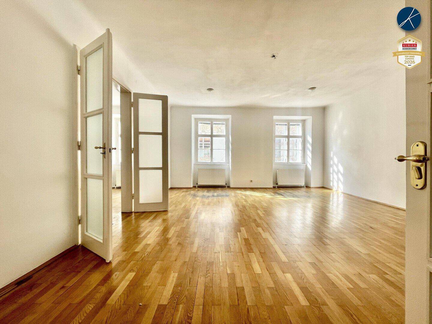Heller Wohnraum mit klassischem Parkettboden und doppelten Flügeltüren, die zu einem weiteren Zimmer führen.