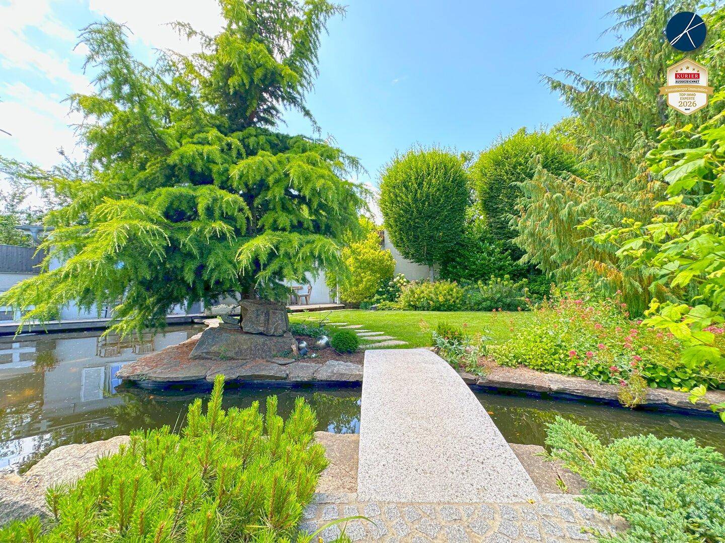 Ruhiger Gartenteich mit kleiner Brücke, umgeben von Bäumen und Sträuchern.