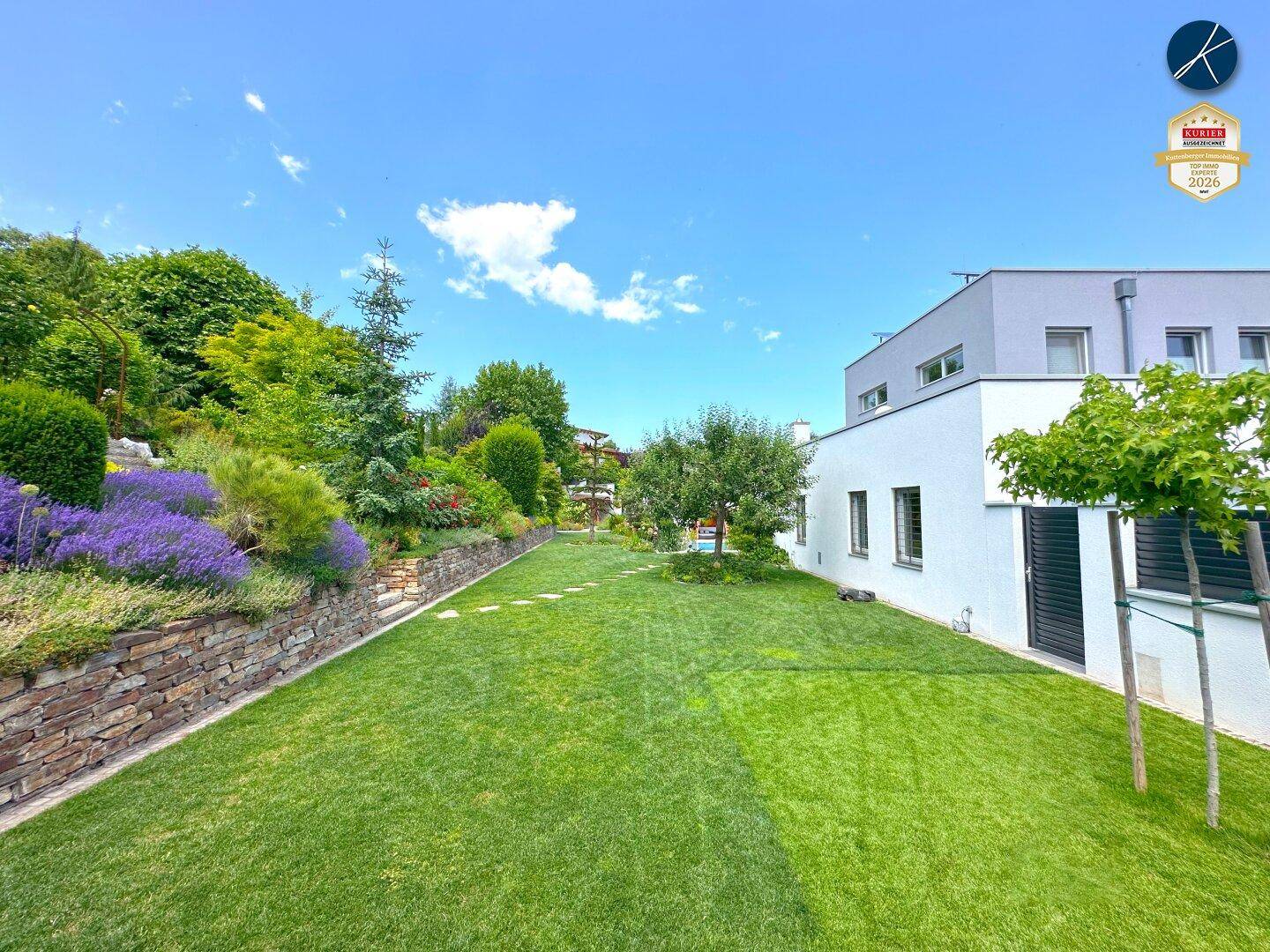 Großer, sonniger Garten mit Rasenfläche, Steinmauer und moderner Hausfassade.