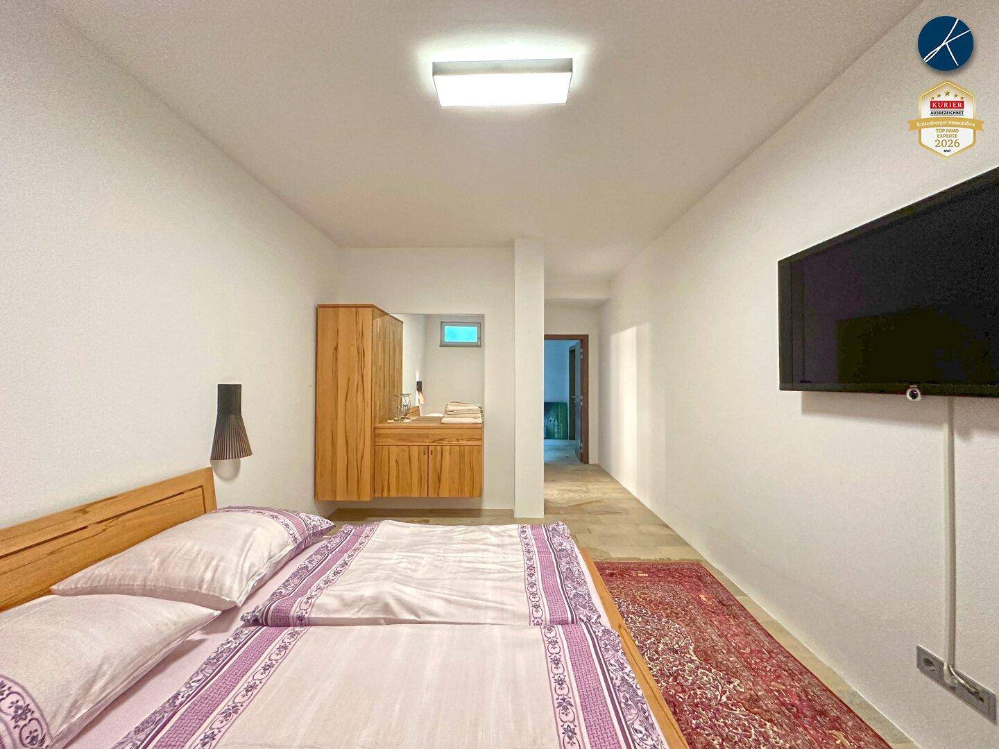 Schlafzimmer mit Doppelbett, Wandfernseher und offenem Zugang zu einem Waschtischbereich.
