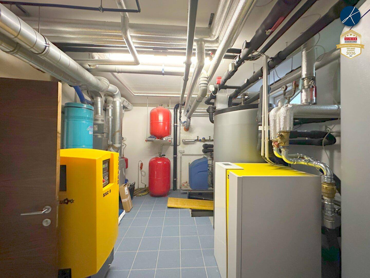 Technikraum mit Heizungsanlagen, Rohren und verschiedenen Speichertanks auf Fliesenboden.