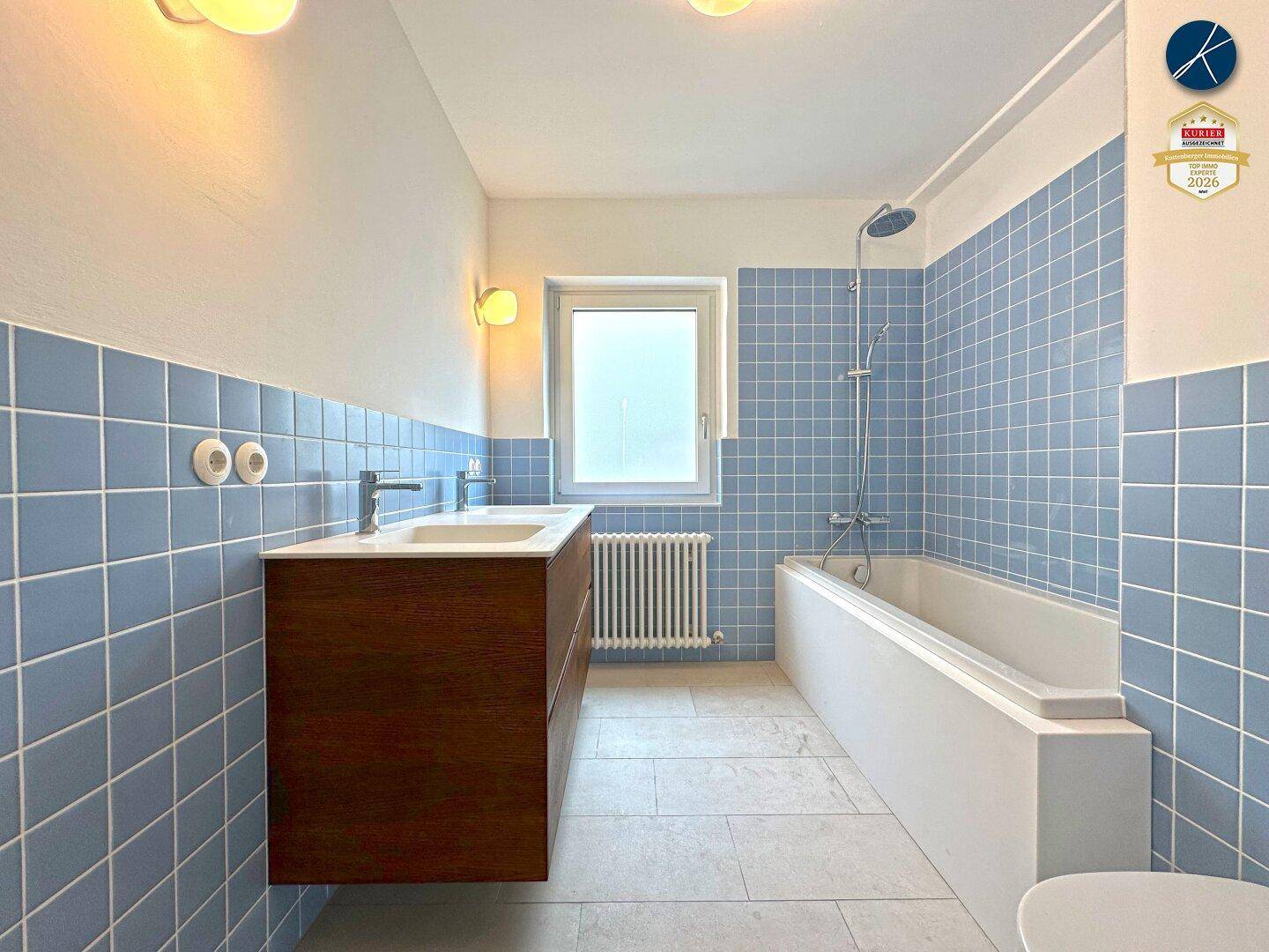 Modernes Badezimmer mit blauen Fliesen, Badewanne mit Duschfunktion, Doppelwaschbecken und Fenster.
