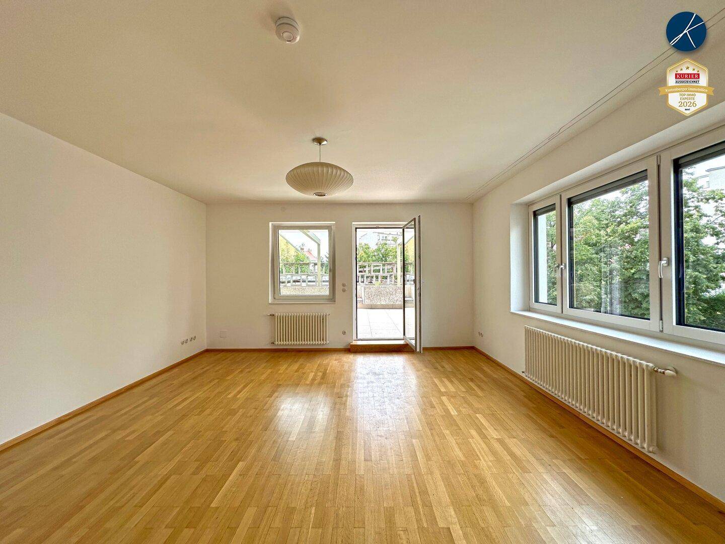 Heller, leerer Raum mit Holzboden und mehreren Fenstern, die Zugang zur Außenterrasse bieten.