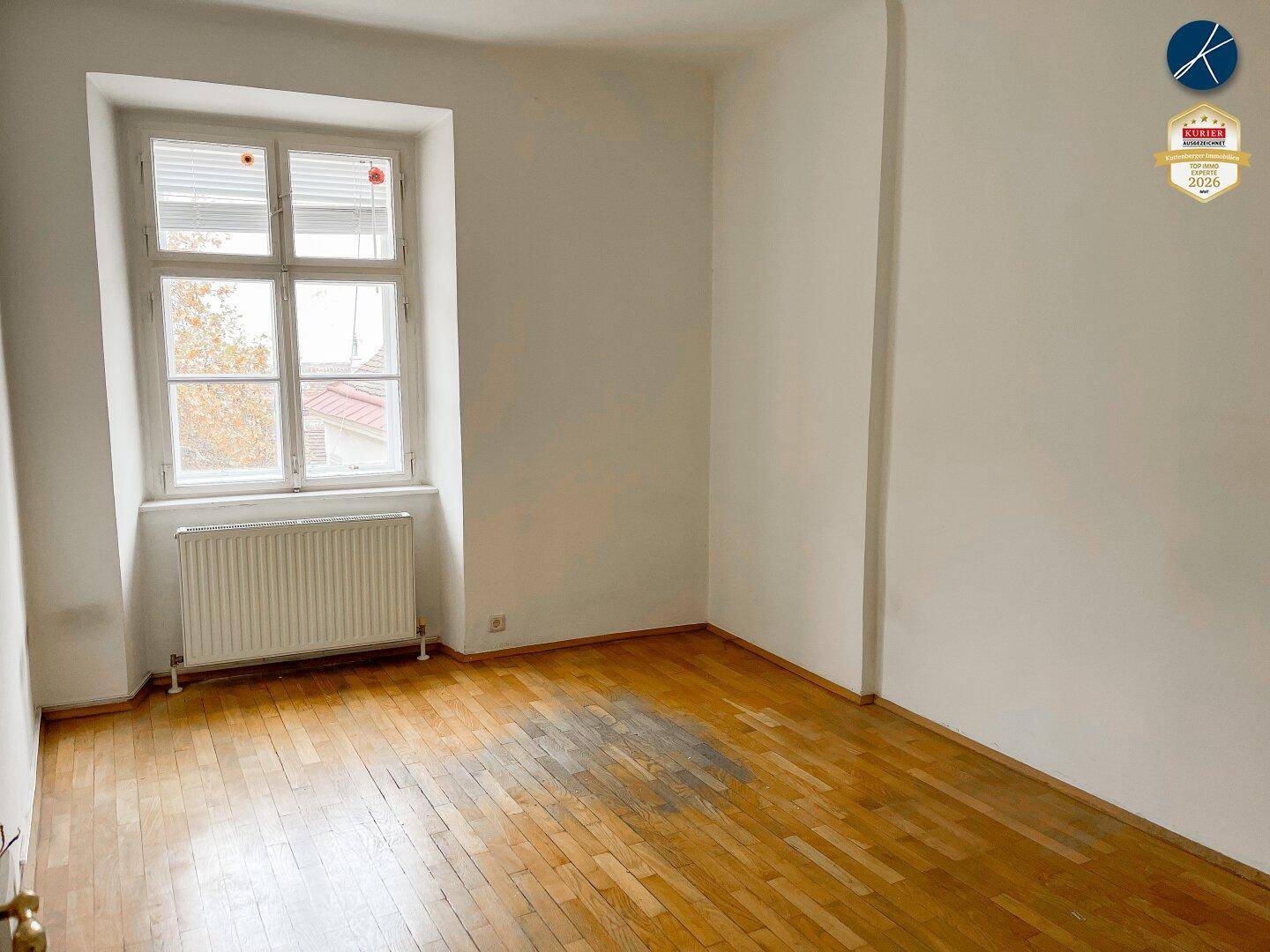 Zimmer mit einem einzelnen Fenster und Parkettboden, der leichte Gebrauchsspuren aufweist.