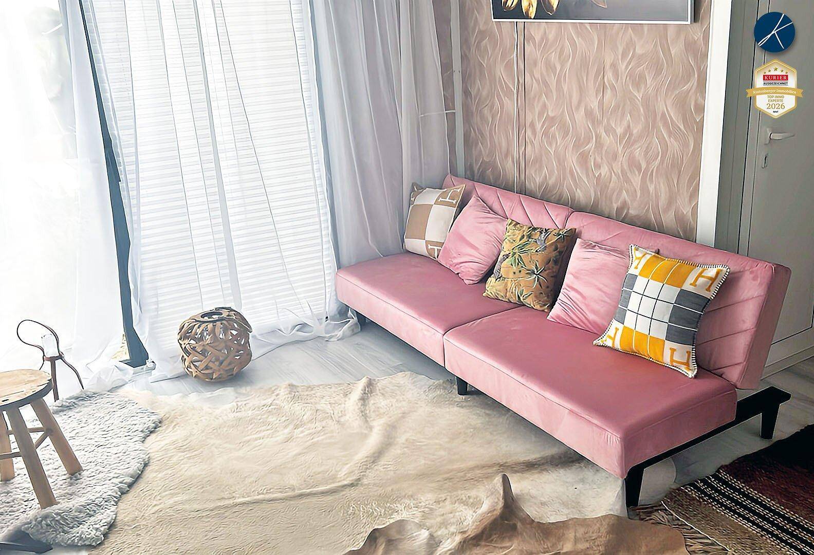 Helles Wohnzimmer mit einem pinkfarbenen Sofa, gemusterter Tapete und großen Fenstern mit Vorhängen.