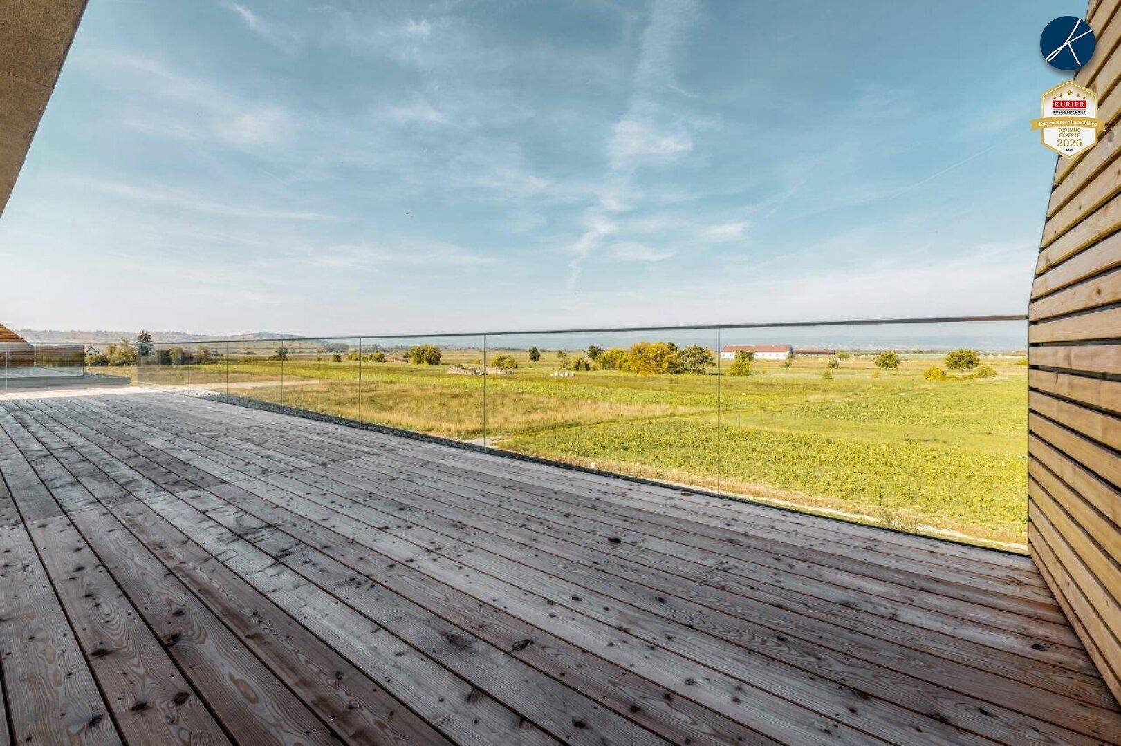 Breite Holzterrasse mit Glasgeländer und unverbautem Blick über die weite, grüne Landschaft.