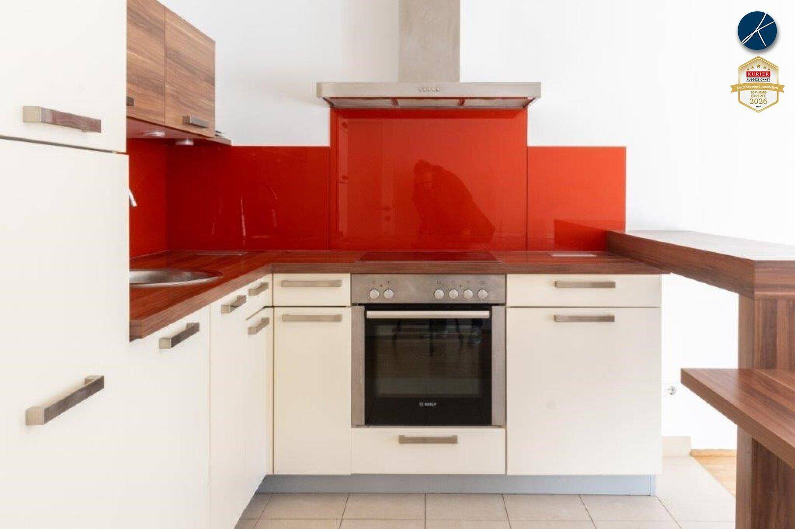 Detailansicht der Küche mit Backofen, Kochfeld und Dunstabzugshaube unter roter Glasrückwand.