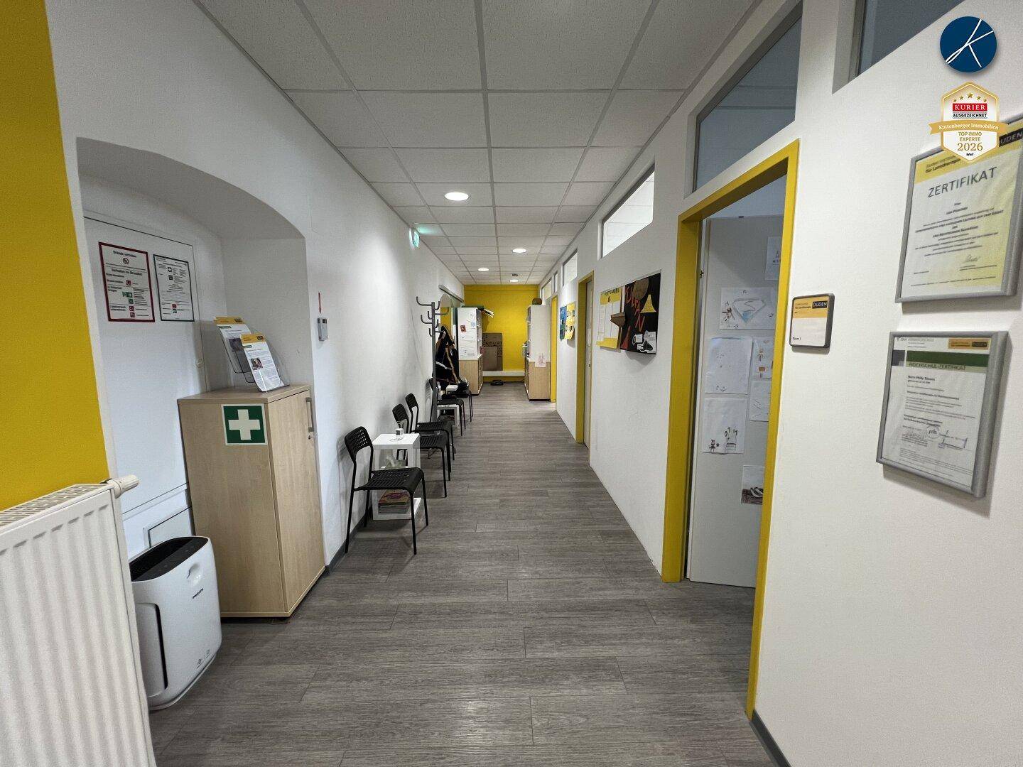 Langer Flur mit gelben Akzentwänden, Wartebereich und mehreren Türen zu Büroräumen.