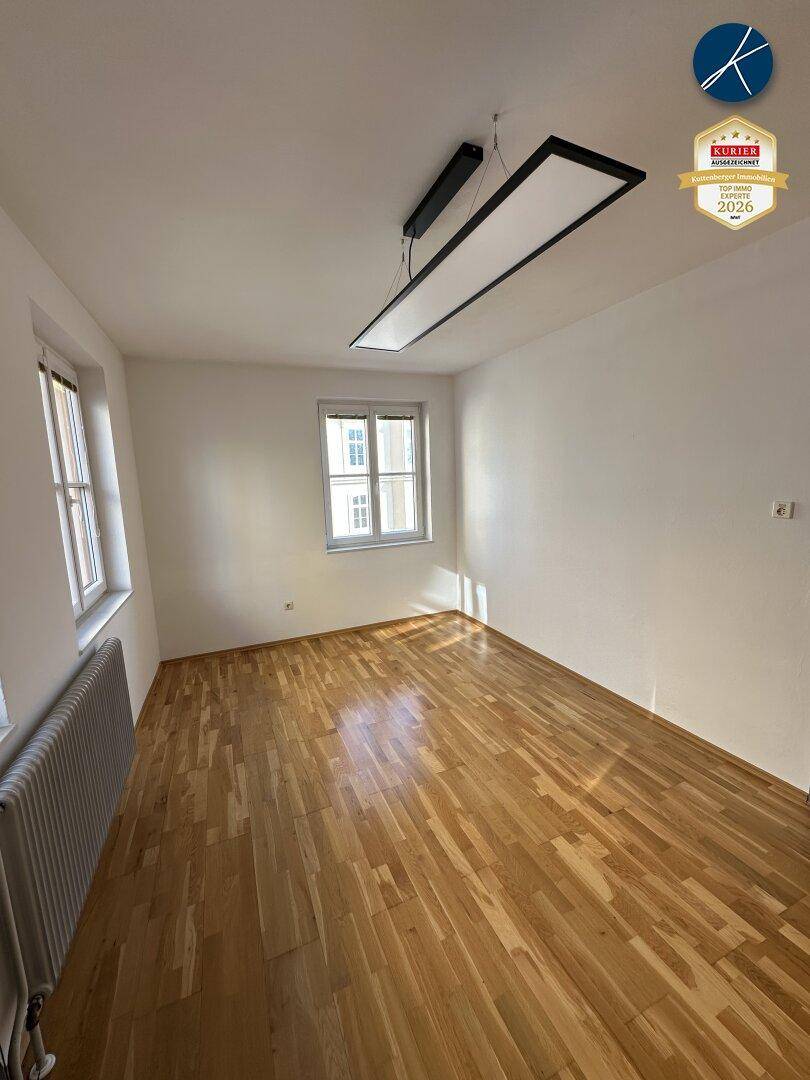 Leeres Zimmer mit Parkettboden und zwei Fenstern, gut belichtet und vielseitig nutzbar.
