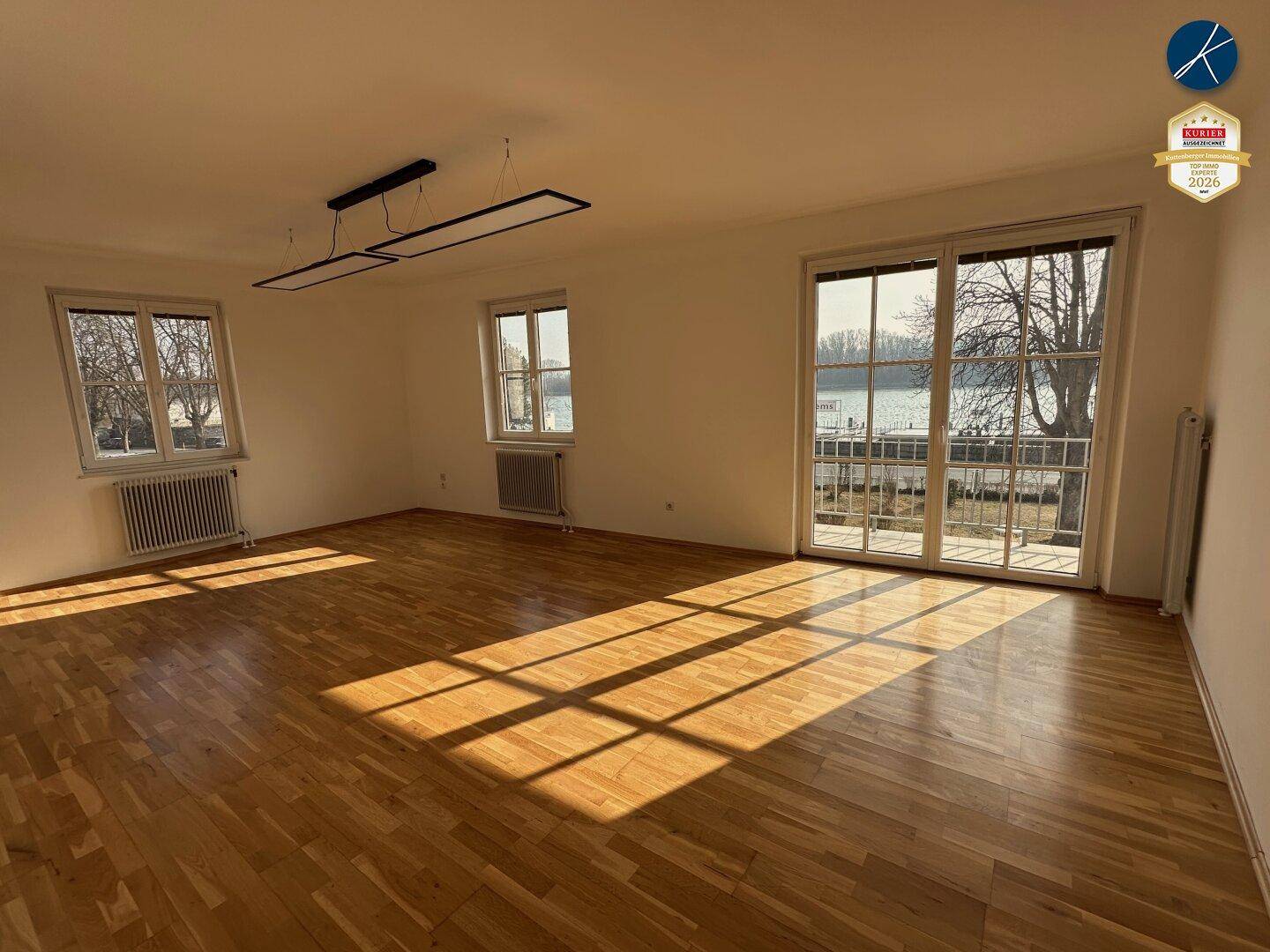 Geräumiges, leeres Wohnzimmer mit hellem Parkettboden, großen Fenstern und viel Tageslicht.