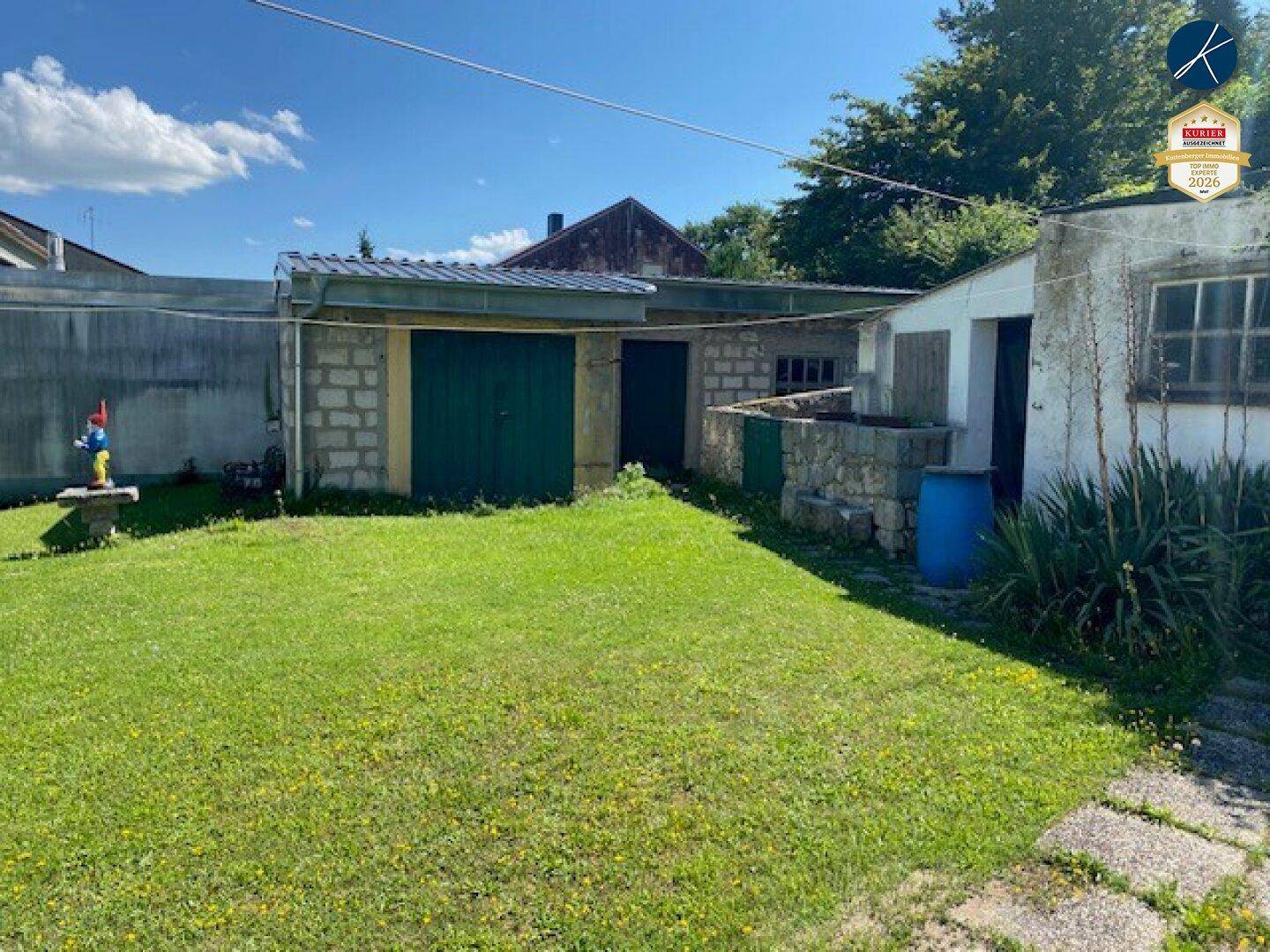 Hinterhofbereich mit Rasenfläche, einfachen Nebengebäuden und einer Garage unter blauem Himmel.