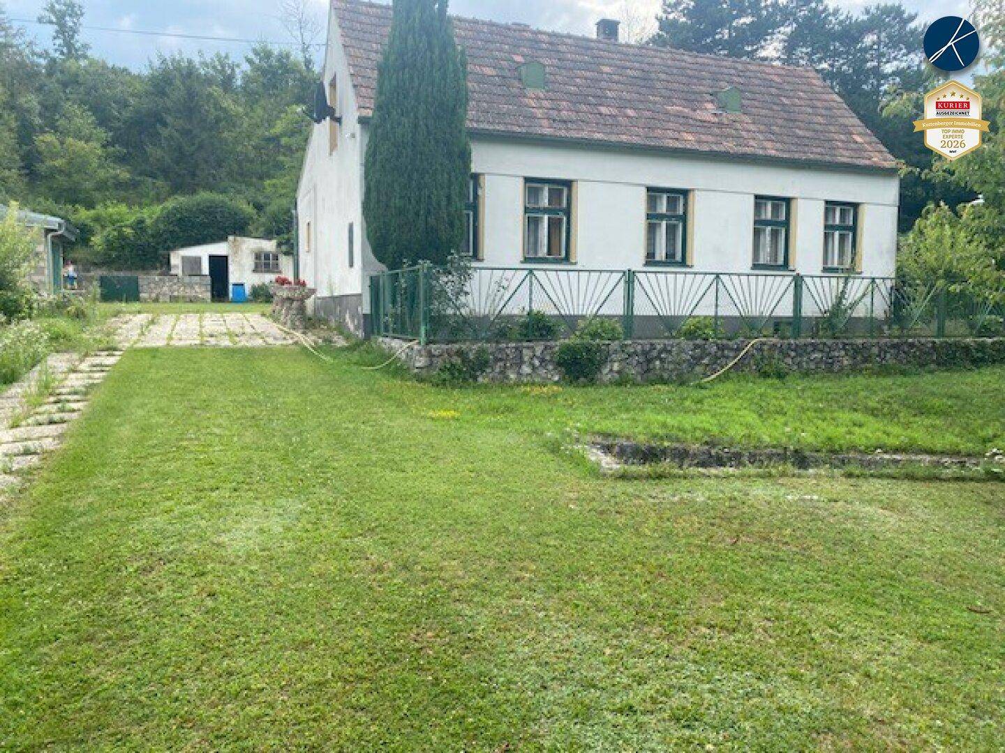 Traditionelles Haus mit weißer Fassade, umgeben von einem grünen Garten und einer Zufahrt unter blauem Himmel.