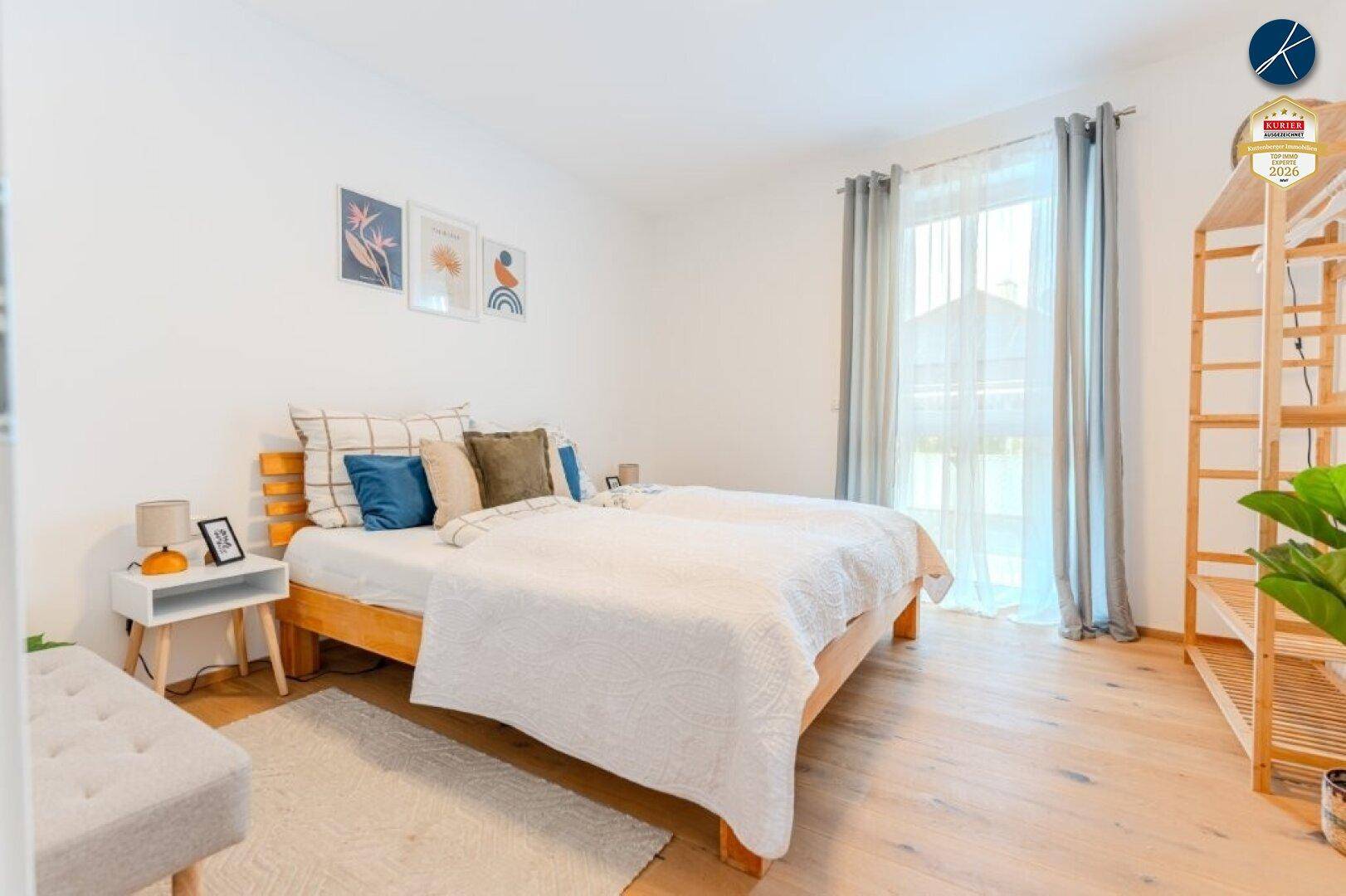 Helles Schlafzimmer mit Holzbett, Nachttisch und großem Fenster mit Vorhängen.