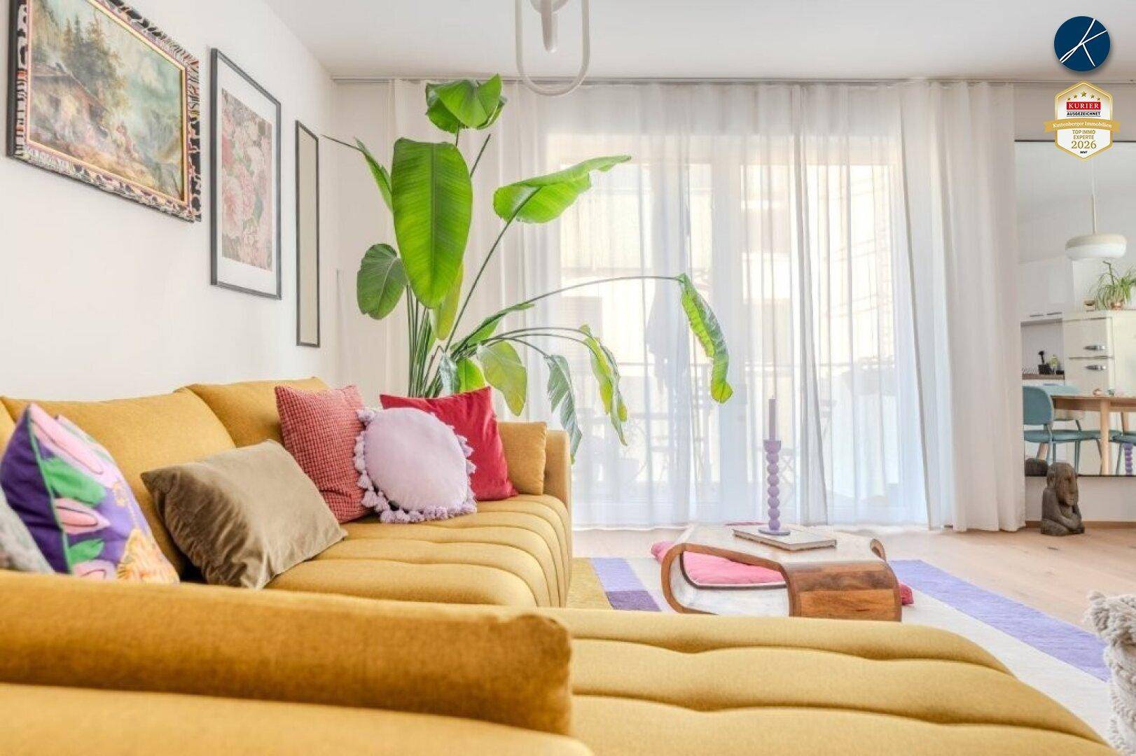 Gemütliches Wohnzimmer mit gelbem Sofa, großen Pflanzen und viel Tageslicht durch die Fenster.