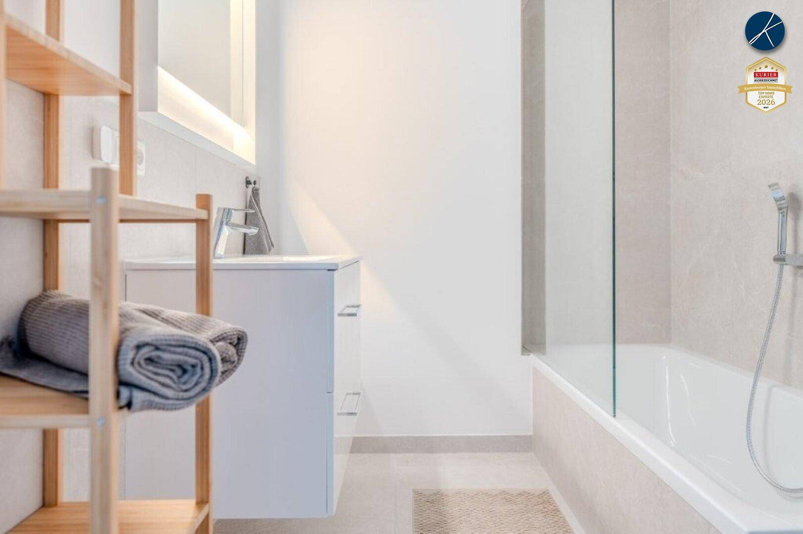 Modernes Badezimmer mit Badewanne, Glasduschwand, weißem Waschtisch und integrierter Spiegelleuchte.