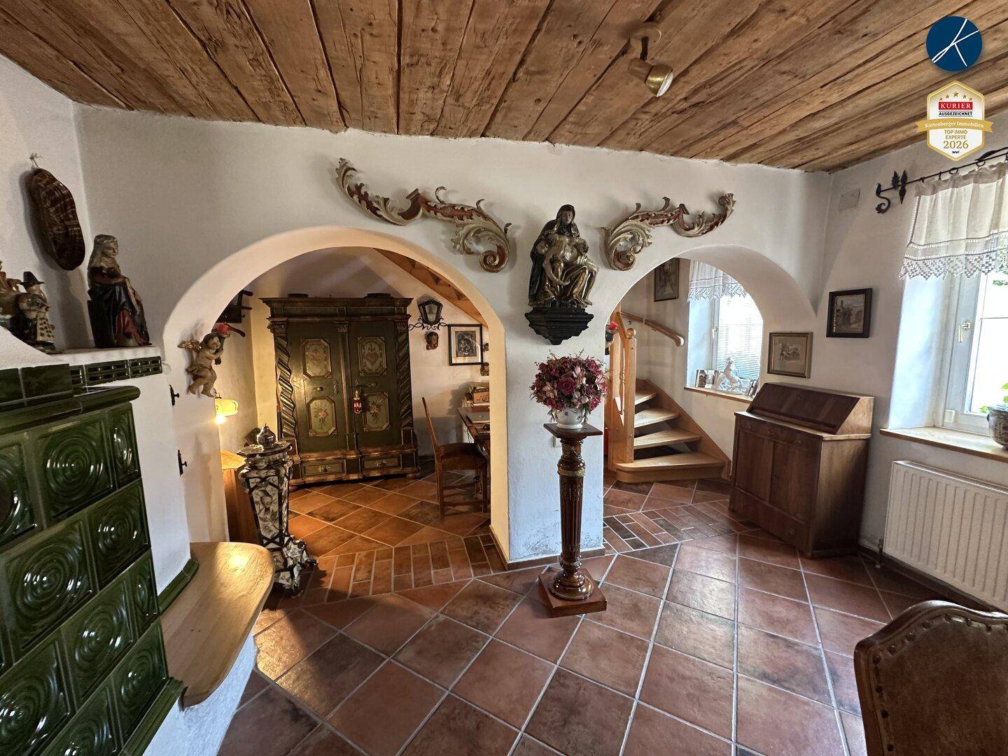 Historischer Eingangsbereich mit Rundbögen, Kachelofen und einer Holztreppe im Hintergrund.