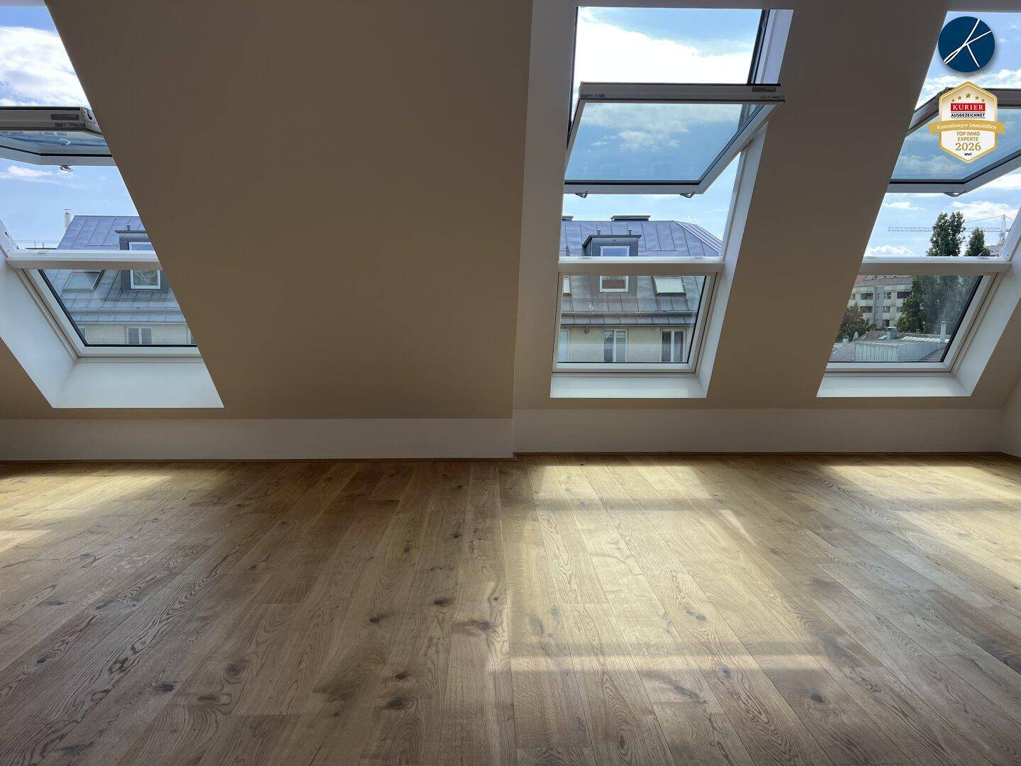 Heller Dachgeschossraum mit Holzboden und Blick auf die umliegenden Dächer durch die Fenster.