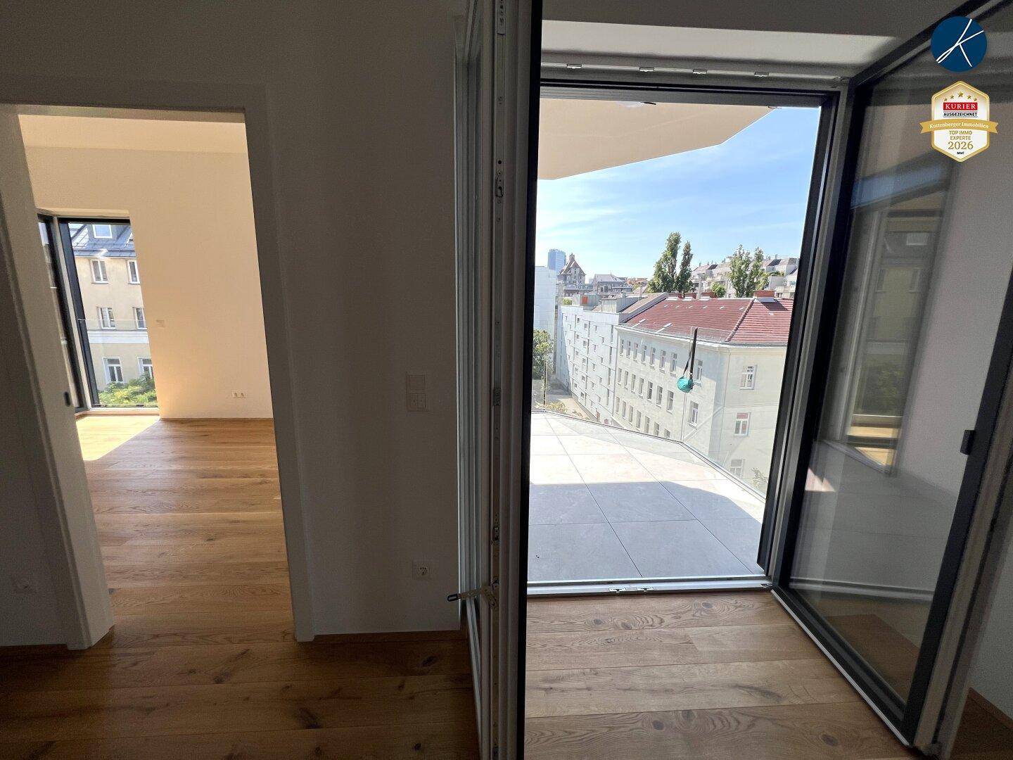 Heller Innenraum mit Holzboden und Zugang zu einem sonnigen Außenbereich mit Stadtblick.