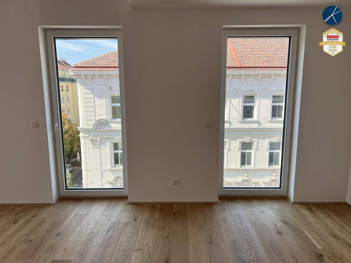 Heller Raum mit Holzboden und zwei großen Fenstern, die einen Blick auf die Stadt bieten.