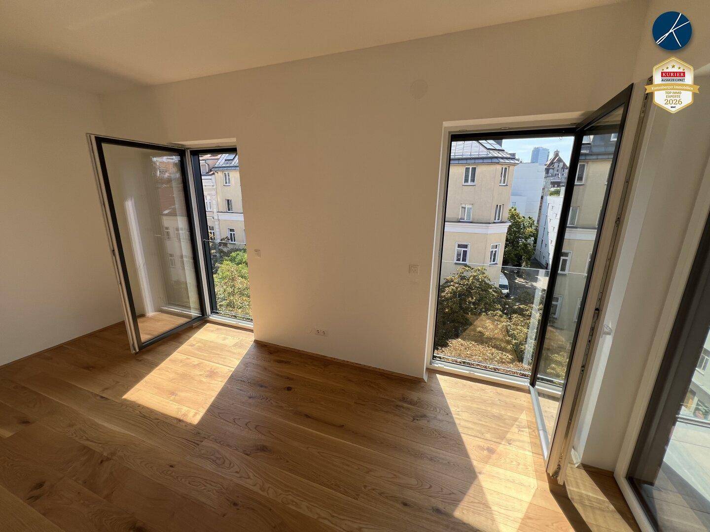 Lichtdurchfluteter Raum mit Holzboden und bodentiefen Fenstern, die einen Stadtblick ermöglichen.