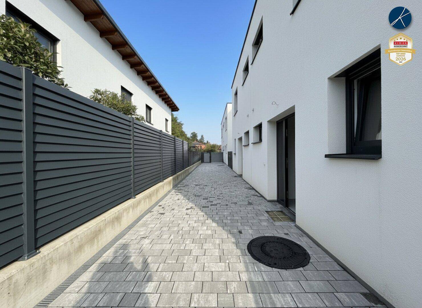Moderner gepflasterter Zugangsweg zwischen zwei weißen Gebäuden mit dunklem Zaun unter blauem Himmel.