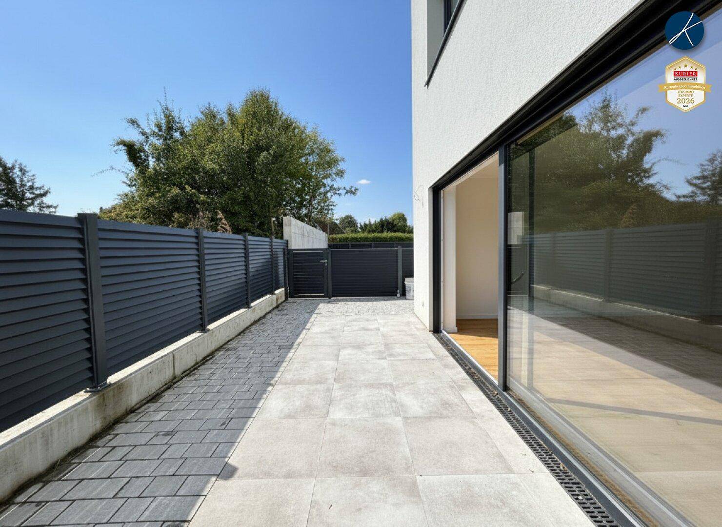 Sonniger Außenbereich mit gepflasterter Terrasse, modernen Glasschiebetüren und einem dunklen Sichtschutzzaun.