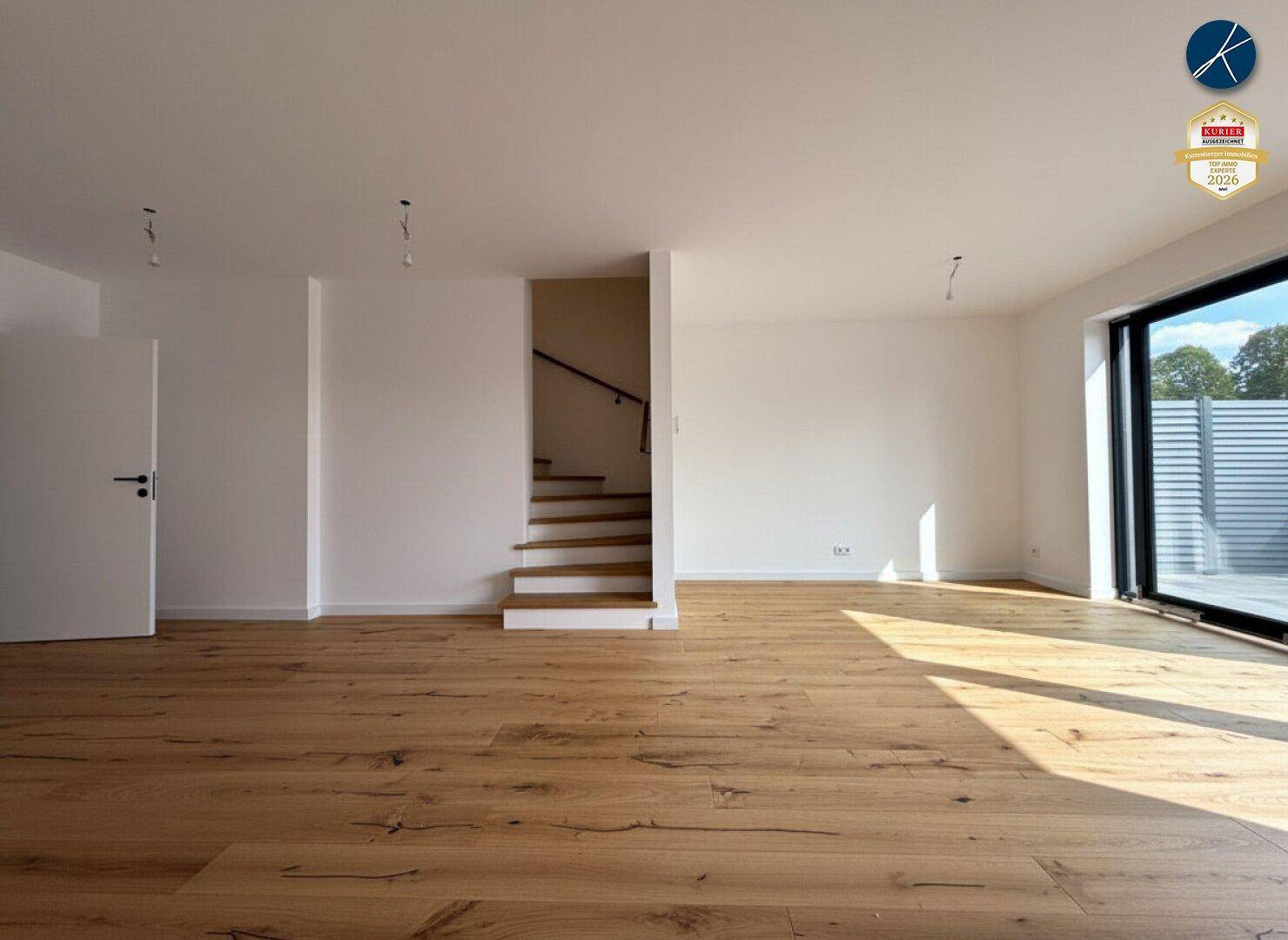 Geräumiges Wohnzimmer mit Holzboden, weiß gestrichenen Wänden und Treppenaufgang.