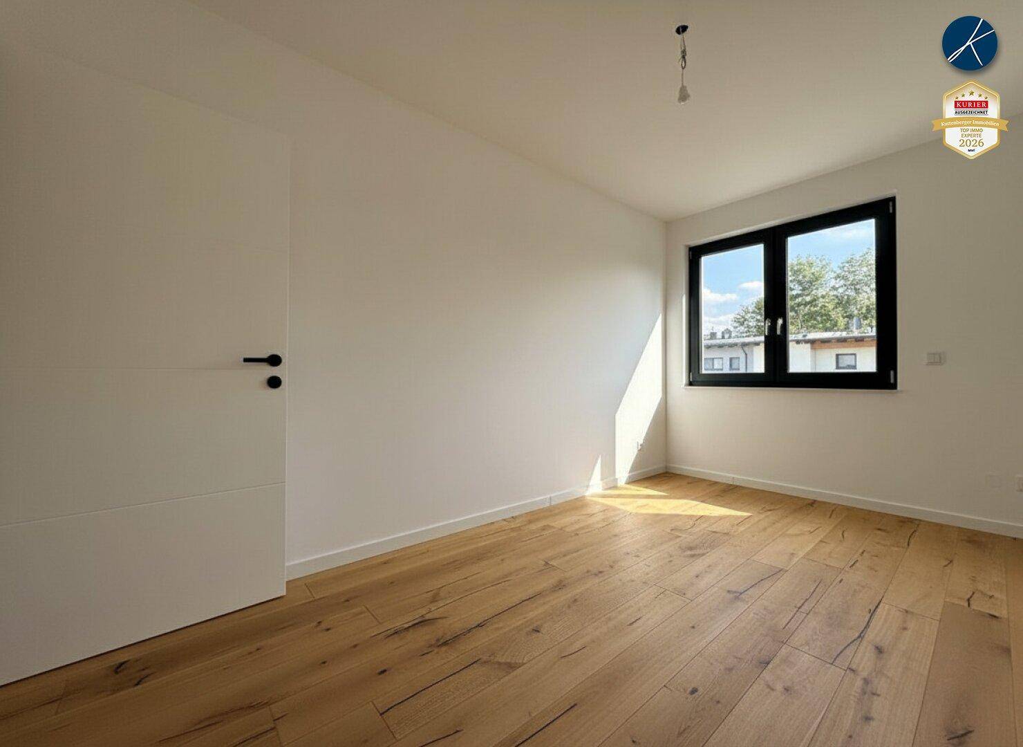 Unmöbliertes Schlafzimmer mit Holzboden, weißer Wandfarbe und Fenster mit Blick ins Grüne.