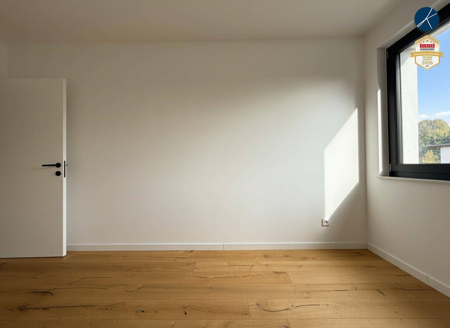 Helles Zimmer mit Holzplankenboden und weißen Wänden, durch das Fenster fällt viel Sonnenlicht.