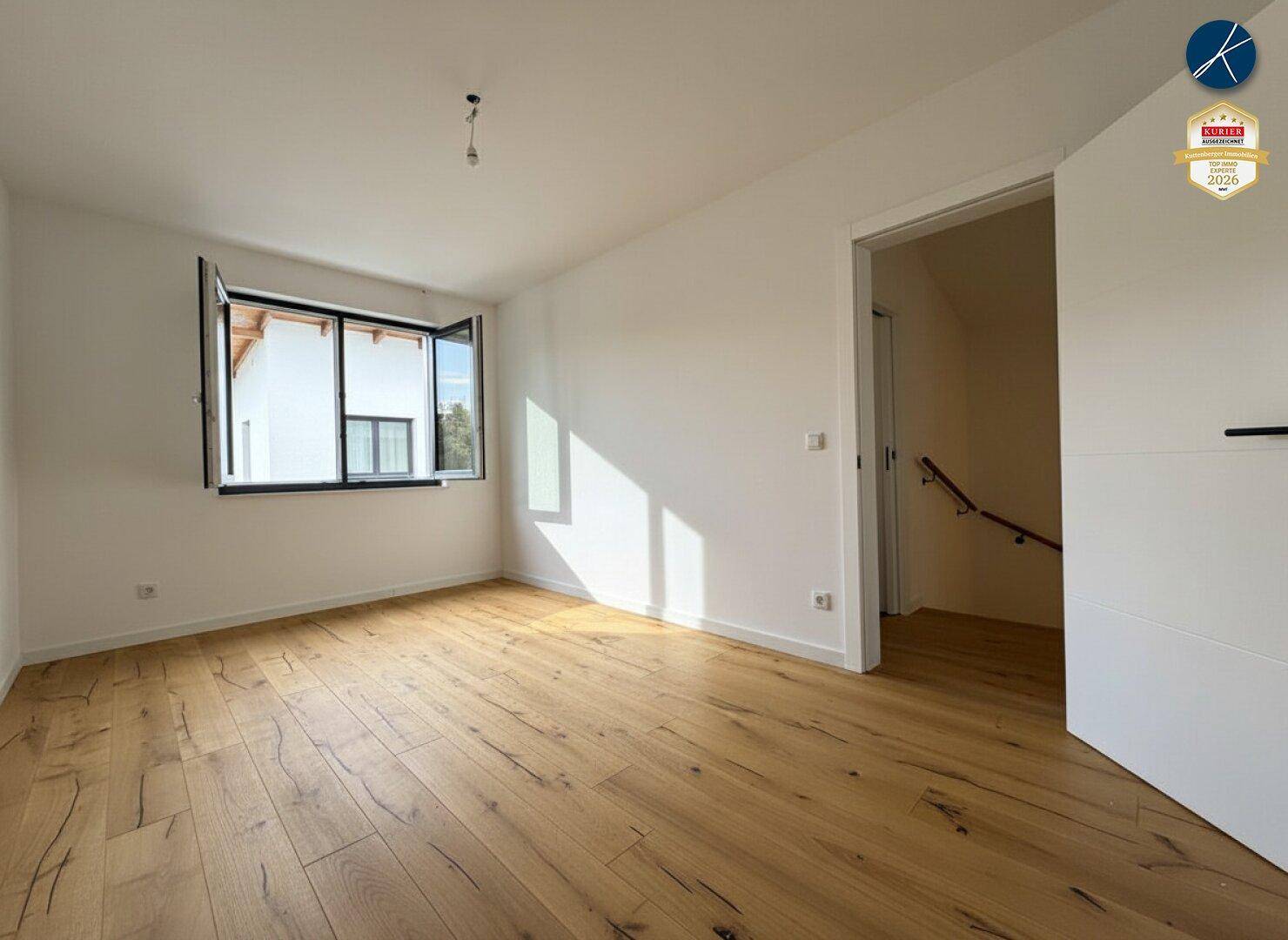 Lichtdurchflutetes Zimmer mit hellem Holzplankenboden, weißen Wänden und Zugang zu einer Treppe.