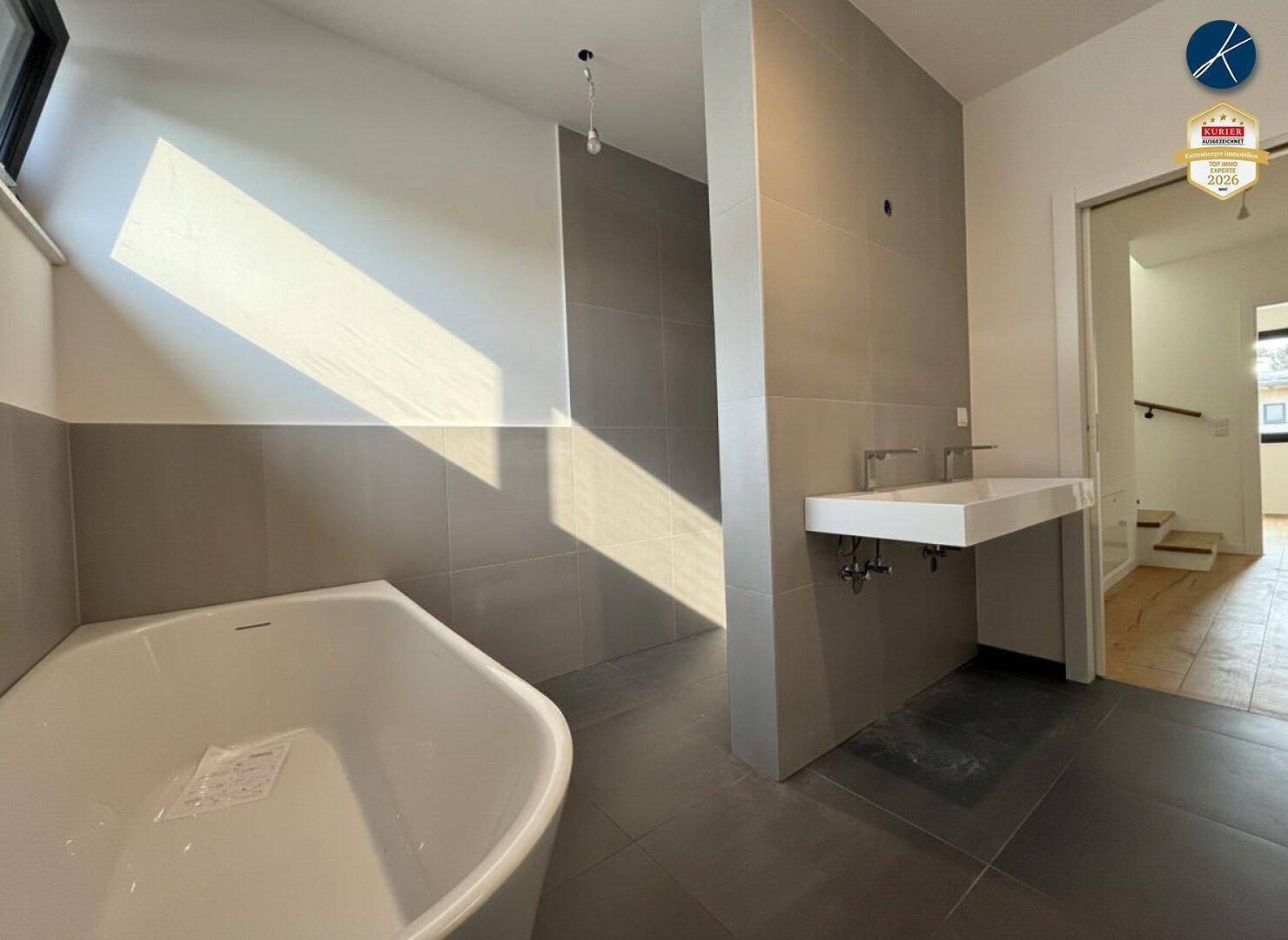 Geräumiges Badezimmer mit moderner Badewanne, Doppelwaschbecken und grauen Fliesen, hell beleuchtet.