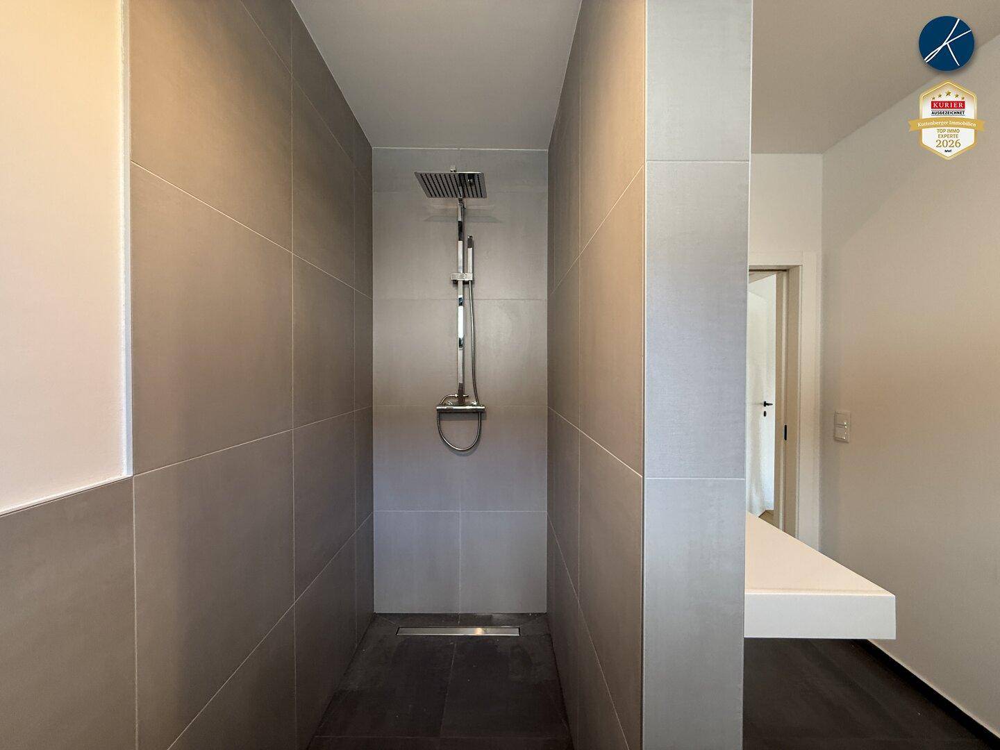 Moderne begehbare Dusche mit Regenduschkopf und großformatigen grauen Fliesen in einem hellen Bad.