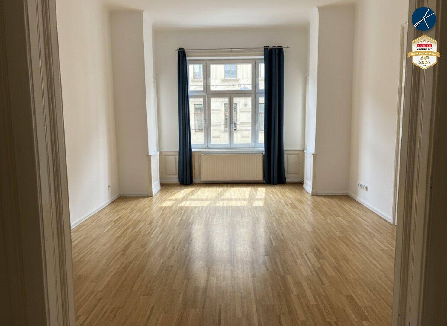 Geräumiges, leeres Zimmer mit Parkettboden und einem großen Fenster mit blauen Vorhängen.