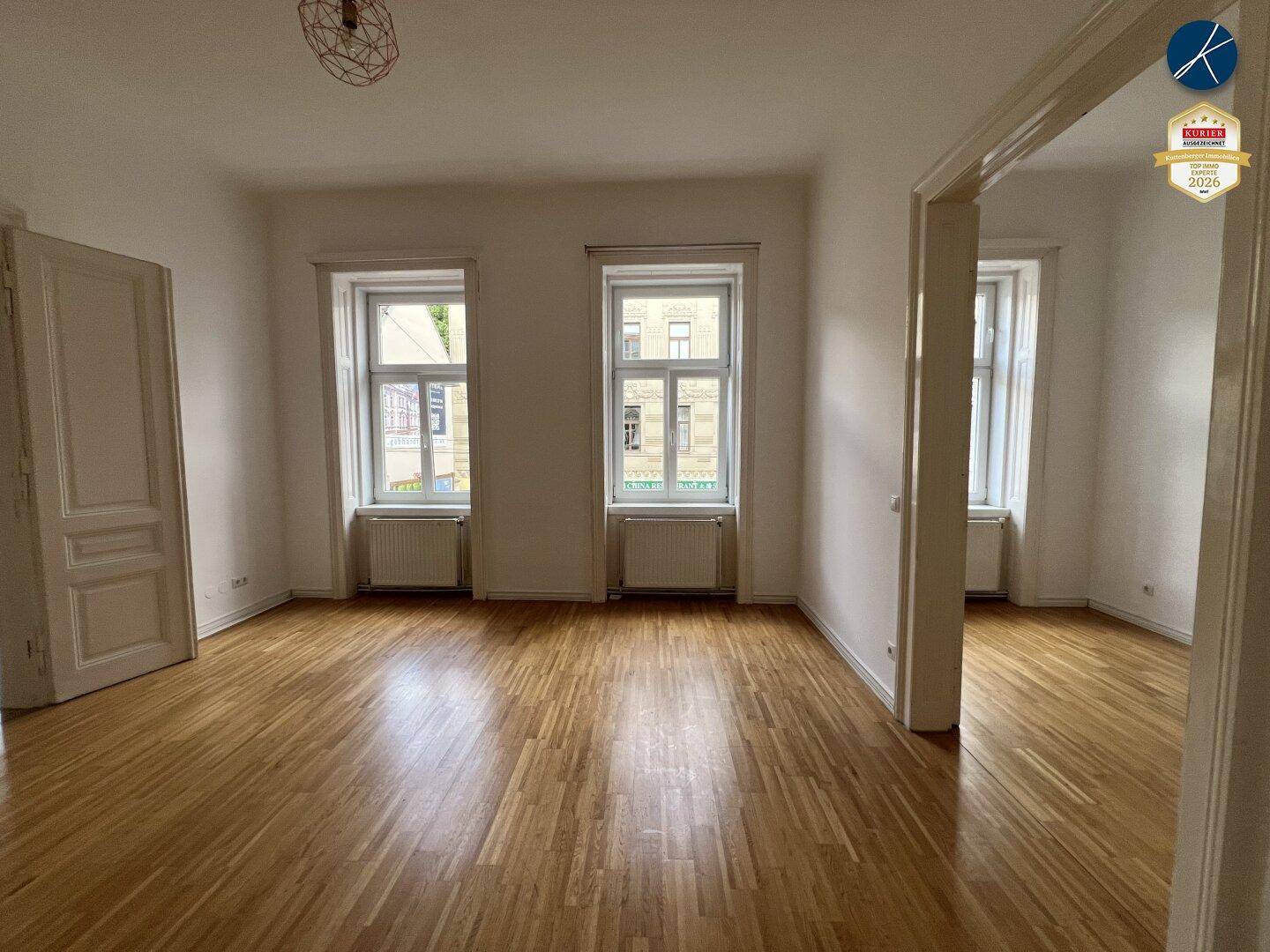 Geräumiges Zimmer mit Parkettboden und zwei großen Fenstern, die viel Licht hereinlassen.