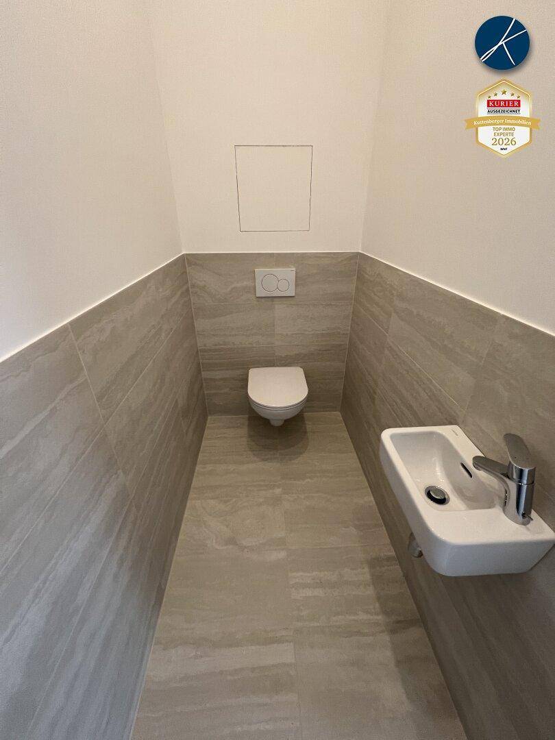 Helles Gäste-WC mit beigefarbenen Fliesen, Wand-WC und kleinem Handwaschbecken.