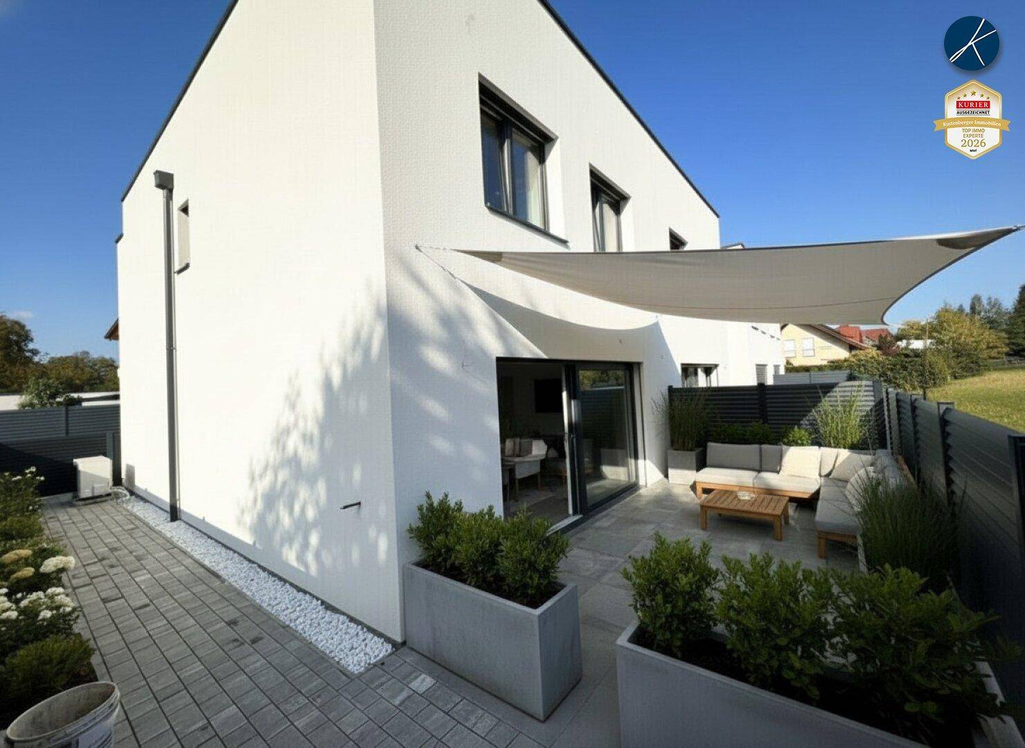 Moderne Terrasse mit Sitzgelegenheiten, Sonnensegel und gepflegten Pflanzenkübeln am Haus.