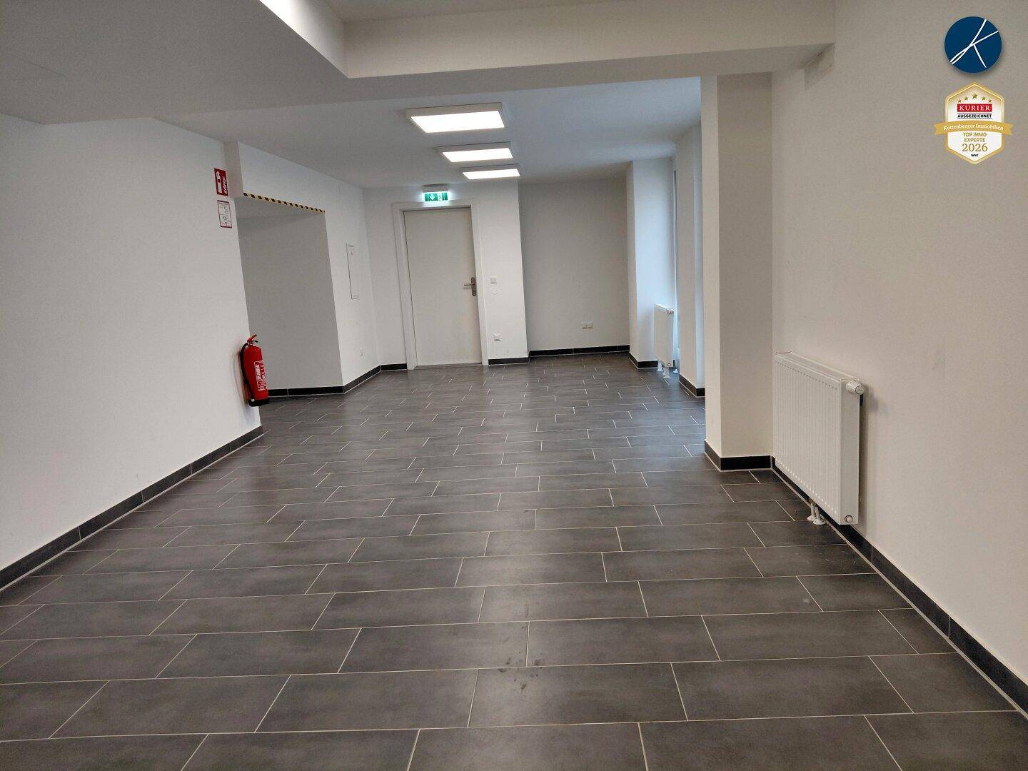 Langer, heller Flur mit dunklem Fliesenboden und weißen Wänden, ausgestattet mit Heizkörpern.