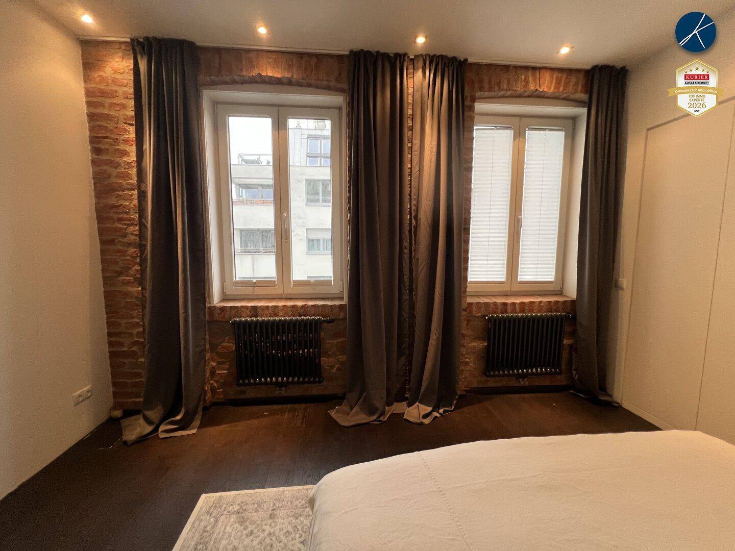 Schlafzimmer mit großem Bett, dunklen Vorhängen und freiliegenden Ziegelwänden.