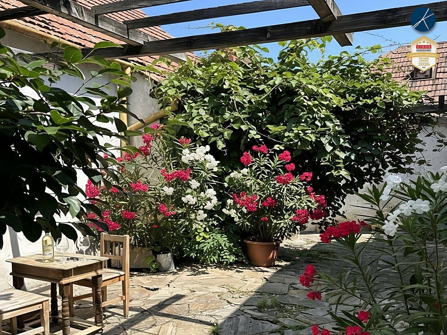 Gemütlicher Außenbereich mit Holztisch und Stühlen, umgeben von üppiger Bepflanzung unter einer Pergola.