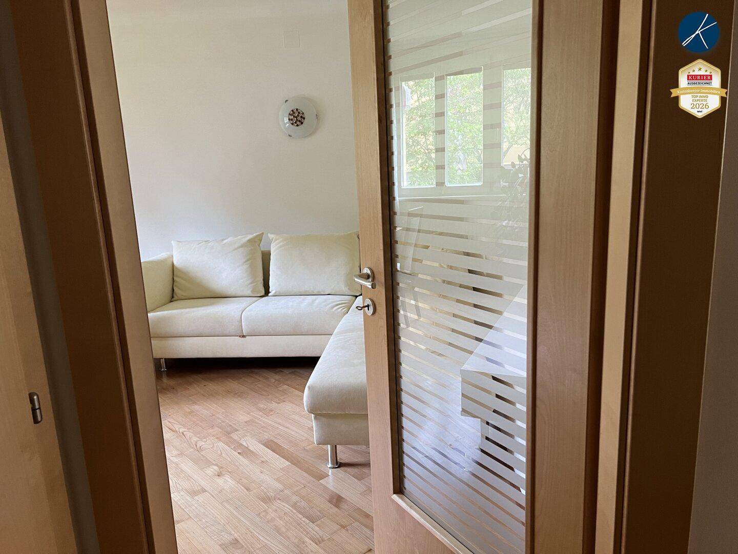 Helles Wohnzimmer mit Holzparkettboden und beigem Sofa, zugänglich durch eine Glastür.